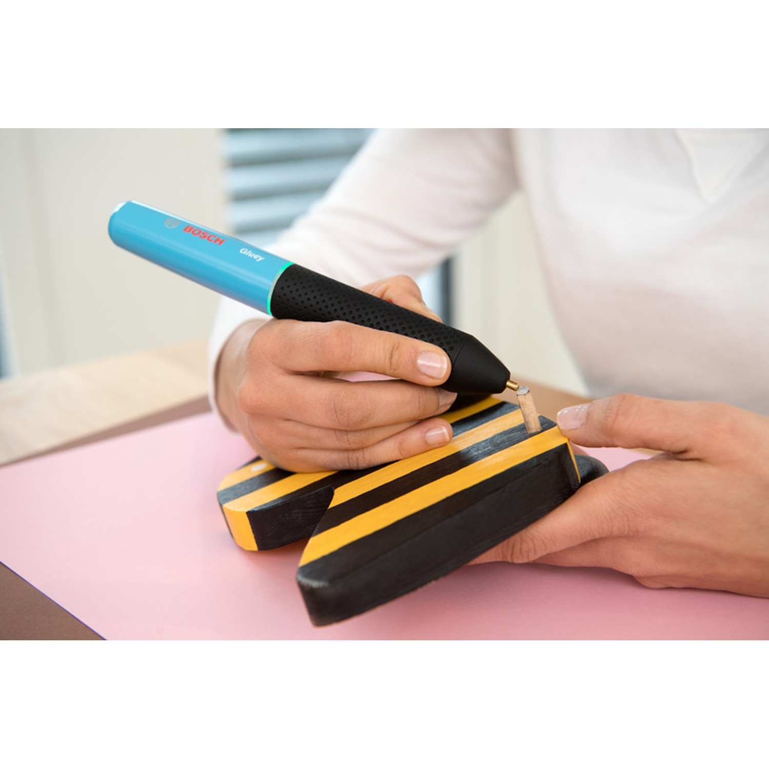 Una persona usa una pistola per colla Bosch blu e nera su un oggetto di legno con strisce gialle e nere, su una superficie rosa.