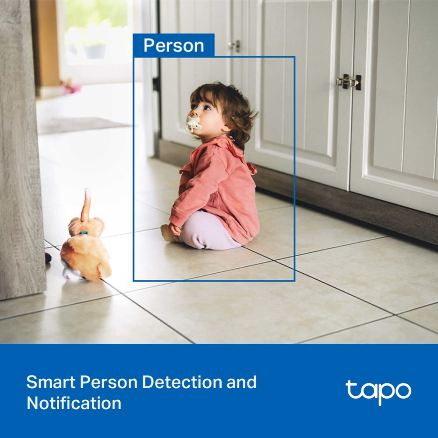 A baby wearing a pacifier sits on a tiled floor. A stuffed toy is in the foreground. Cabinets are in the background.