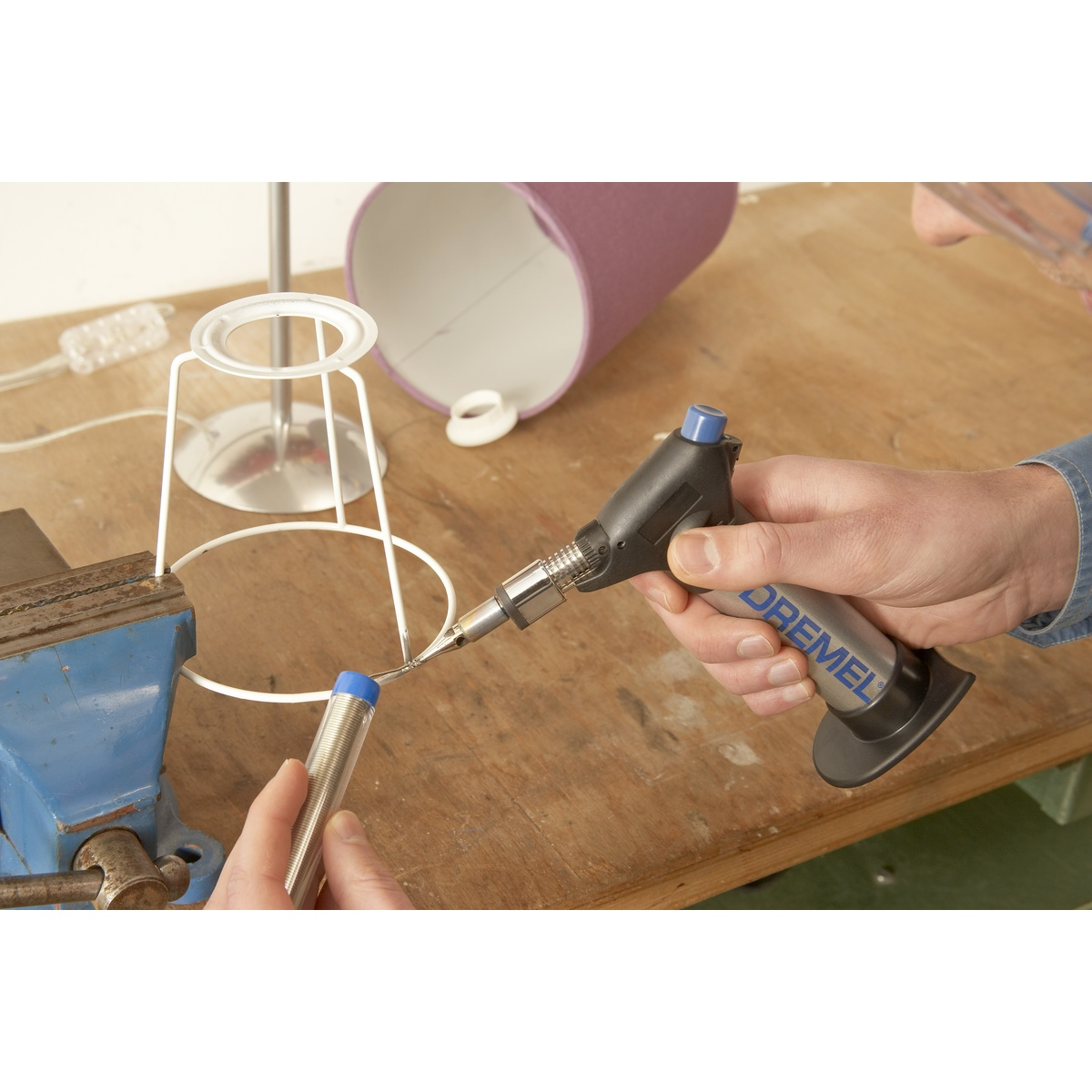 Una persona che salda una base di lampada. Viene utilizzato un utensile Dremel. Lo sfondo mostra un paralume e un tavolo di legno.