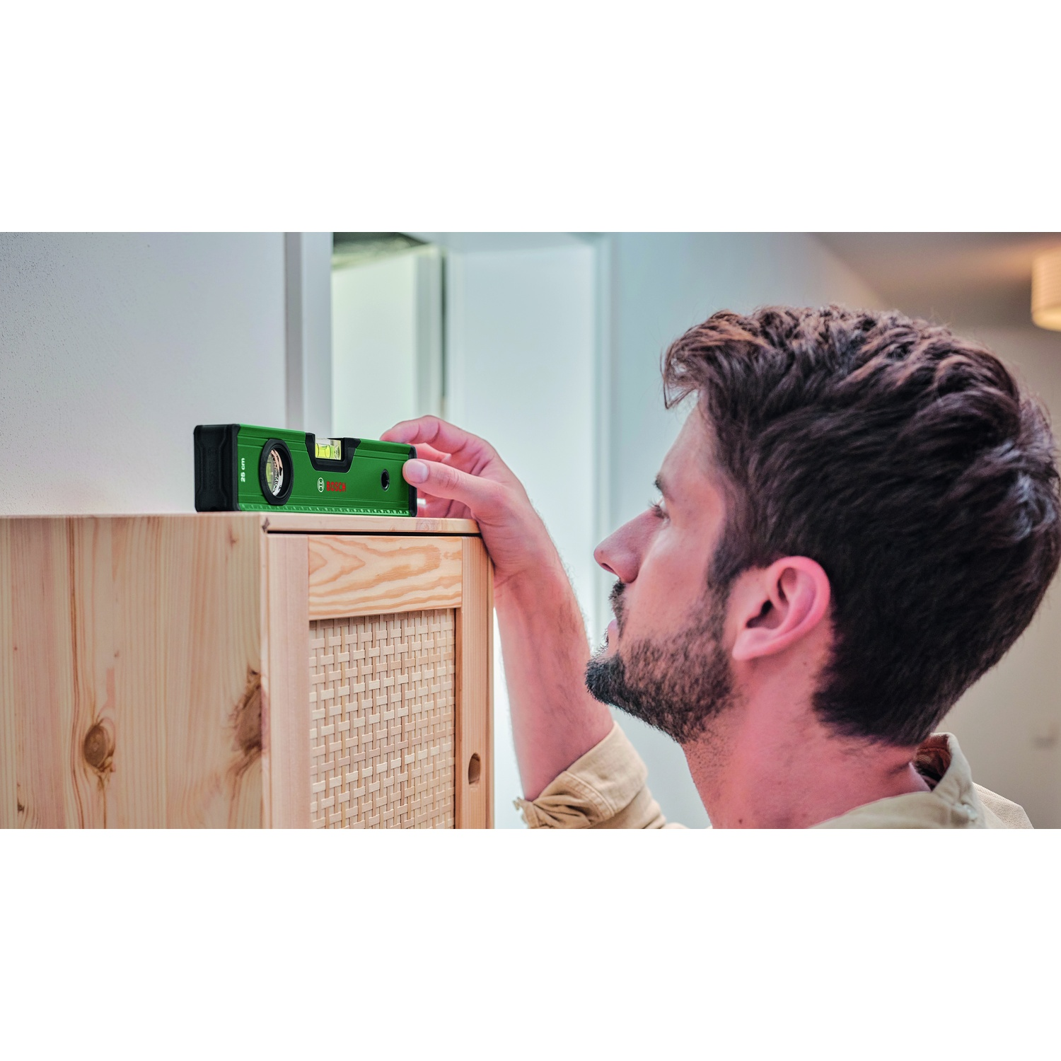 Un uomo usa una livella per controllare un mobile di legno. La livella è verde. L'uomo indossa una camicia beige e ha la barba.