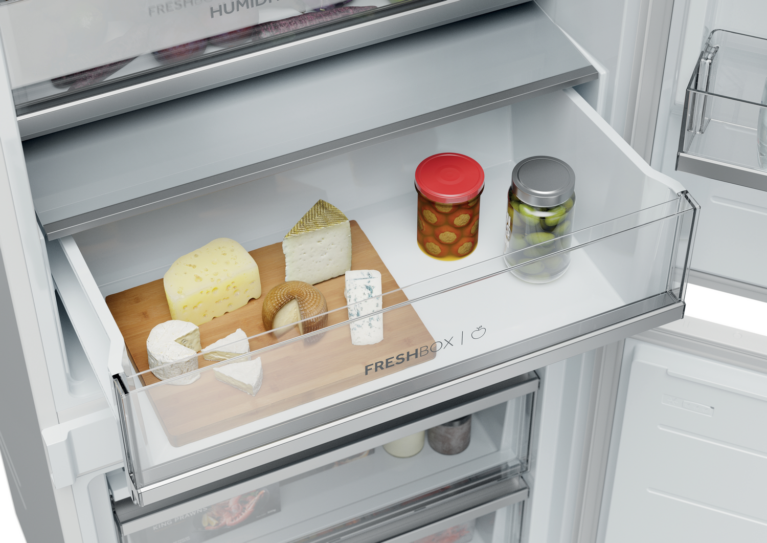 Un cajón de refrigerador contiene queso, frascos de comida y una tabla de madera. El interior es blanco y la puerta está abierta.