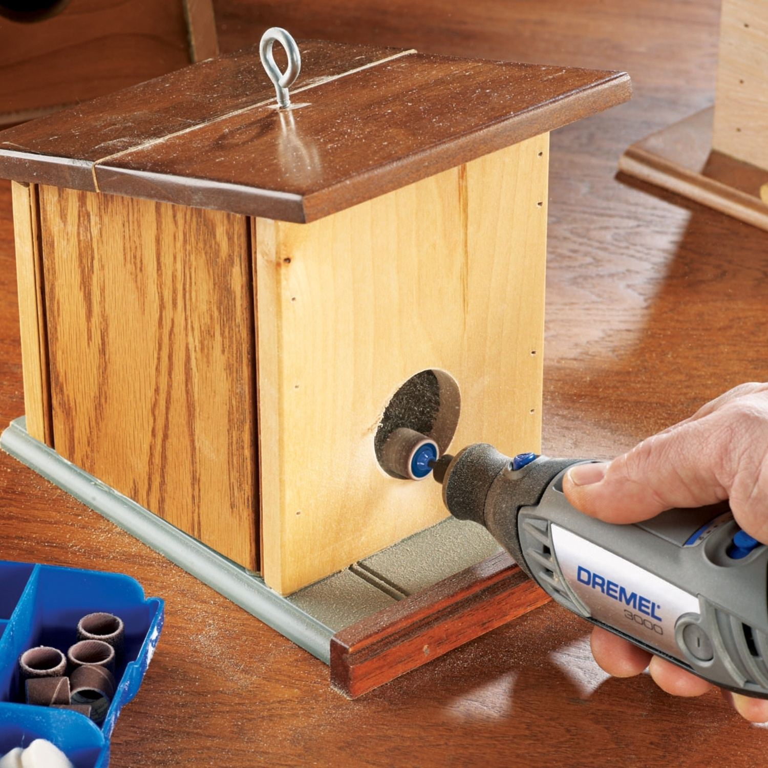 Una persona usa un Dremel per lavorare su una casetta per uccelli di legno. La casetta è marrone chiaro con un tetto marrone scuro. Un vassoio blu per parti è vicino.