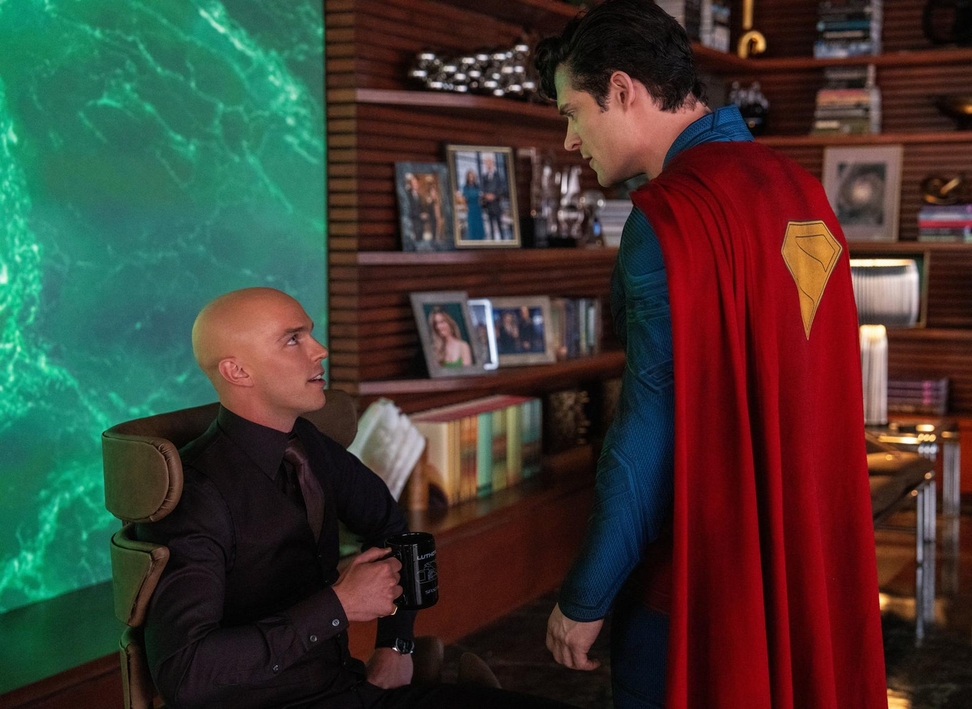Superman steht Lex Luthor gegenüber. Luthor sitzt mit einer Kaffeetasse. Dahinter ein Bücherregal und eine grüne abstrakte Tafel.
