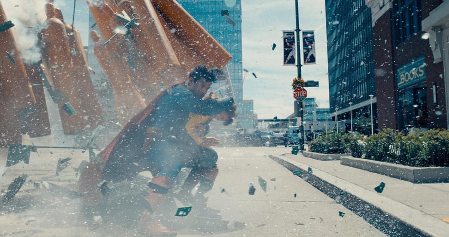 Superman und eine andere Person inmitten einer Trümmer gefüllten Explosion. Gebäude und eine Straße mit Stoppschild im Hintergrund.
