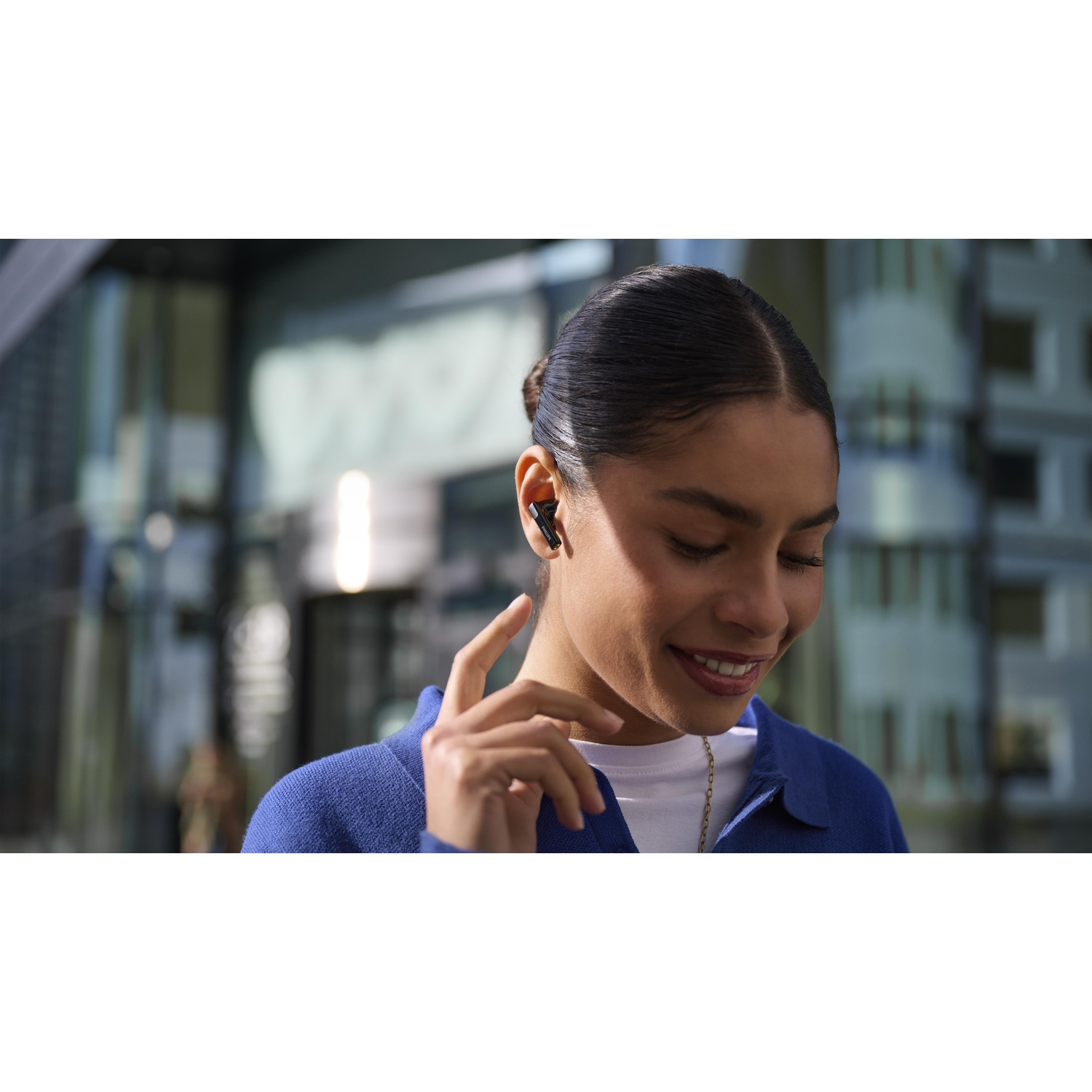 Una donna sorride, indossando auricolari. Si tocca l'orecchio. Gli edifici sono sullo sfondo, la donna indossa una giacca blu e una camicia bianca.
