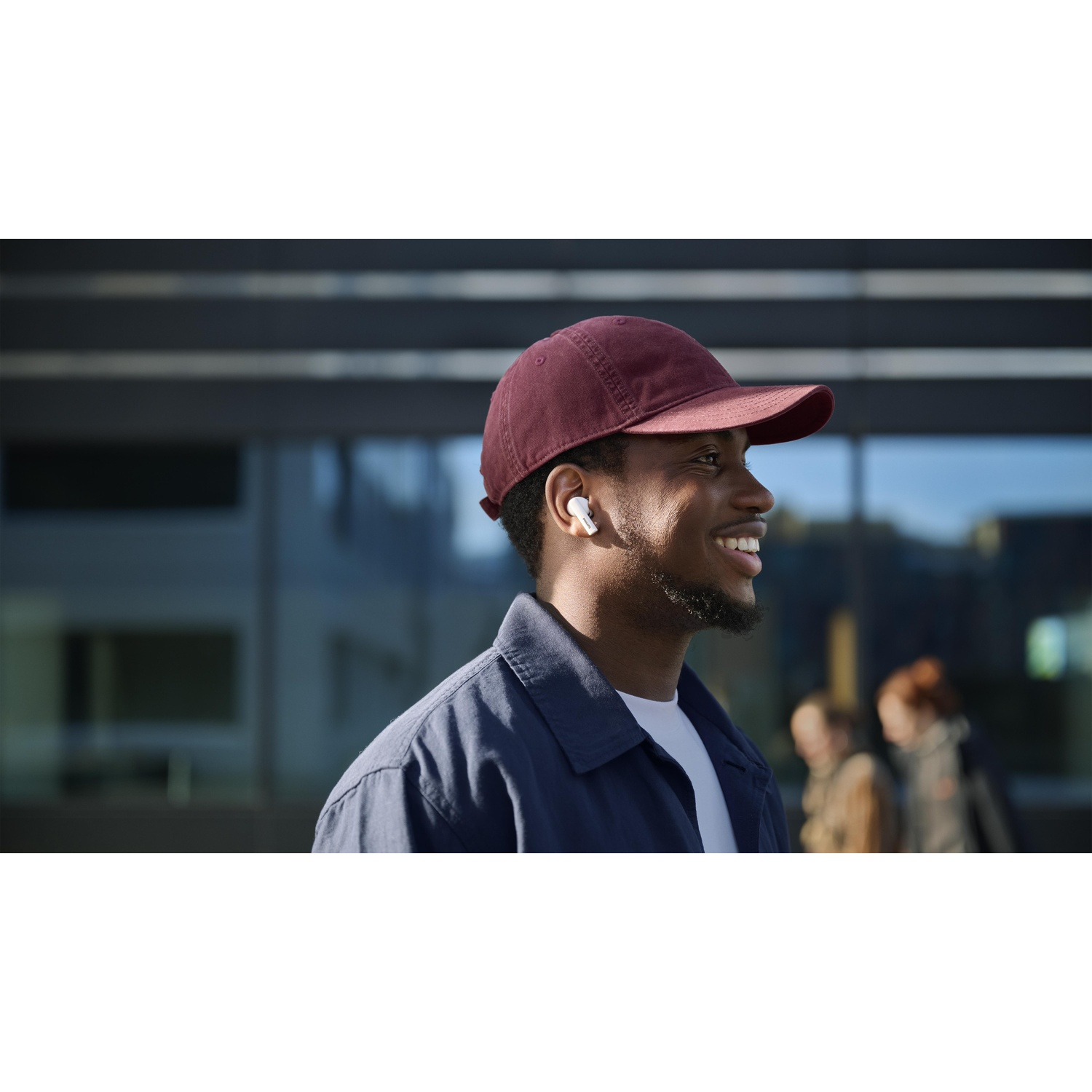 Uomo nero sorridente con berretto bordeaux e auricolari. Indossa una giacca blu e una camicia bianca. Sfondo sfocato.