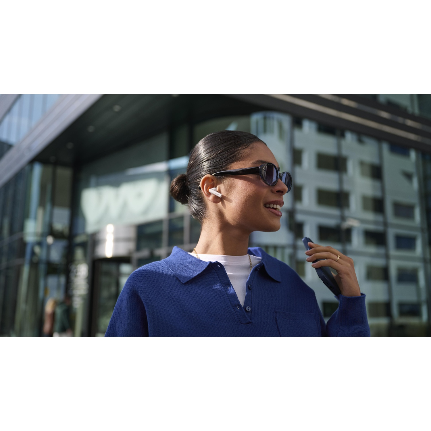 Donna con occhiali da sole e auricolari sorride, tenendo un telefono. Indossa una camicia blu, davanti a un edificio moderno.