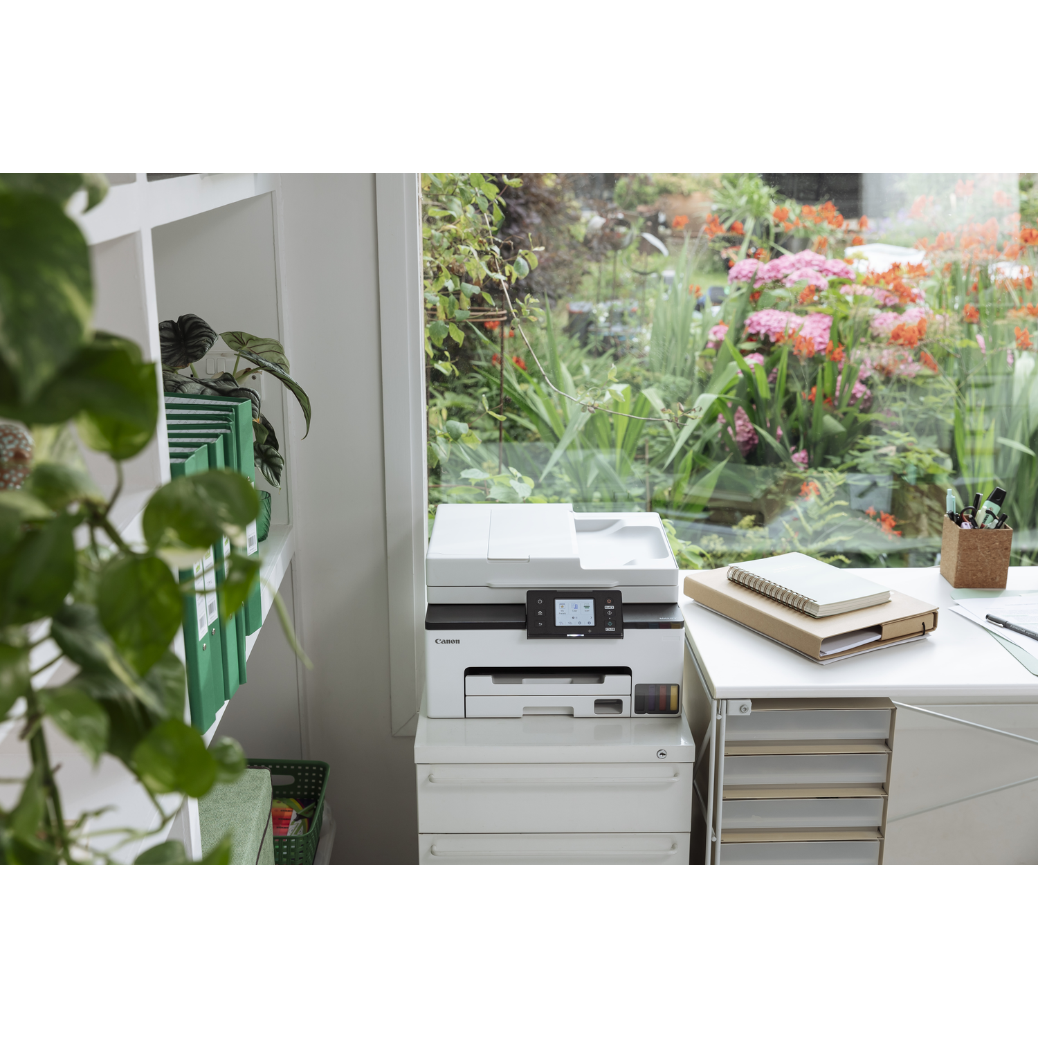 A white Canon printer sits on drawers near a window. A desk has notebooks, pens, and a view of a garden with flowers.