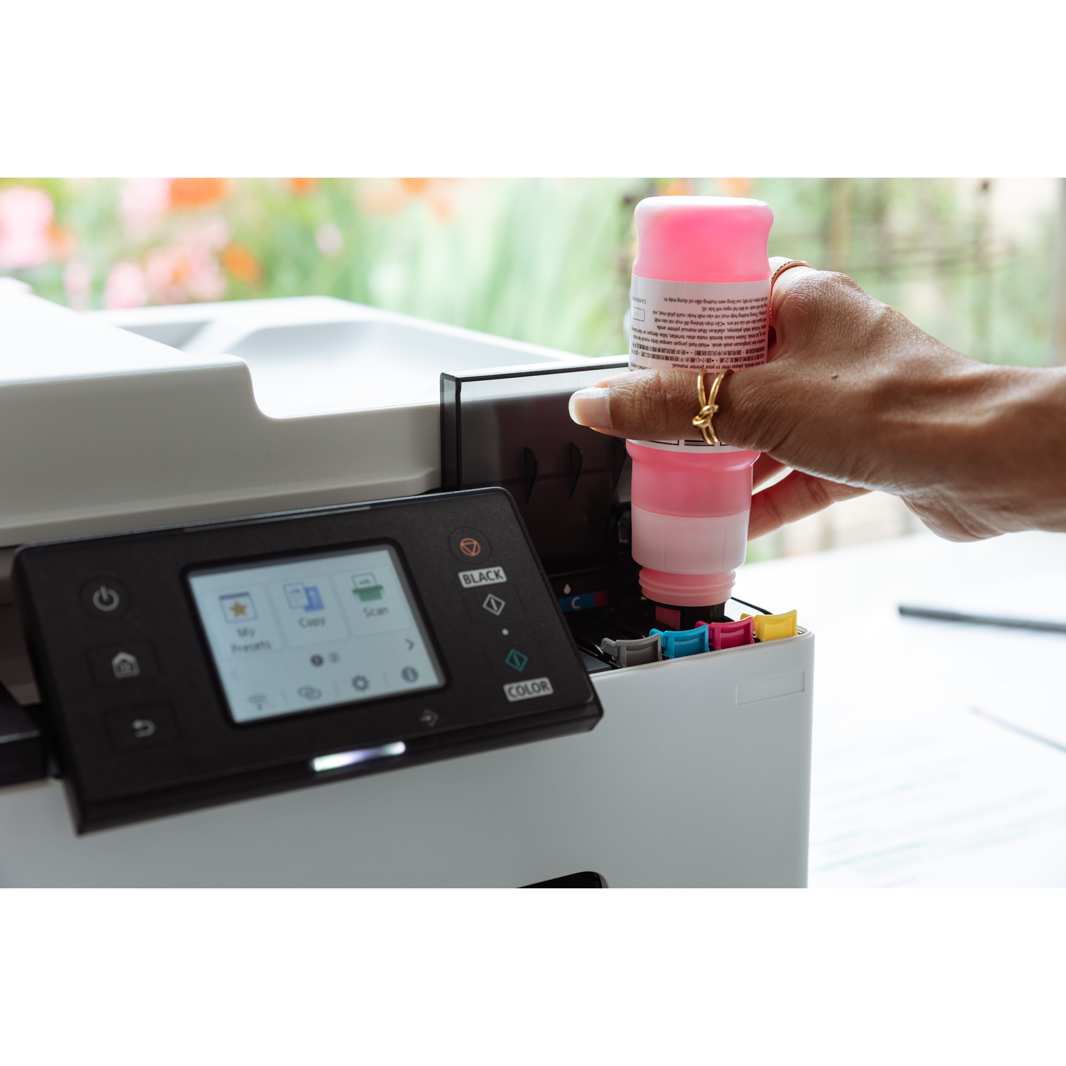 A hand inserts a pink ink cartridge into a printer. The printer has a display showing options. Background shows a blurred garden.