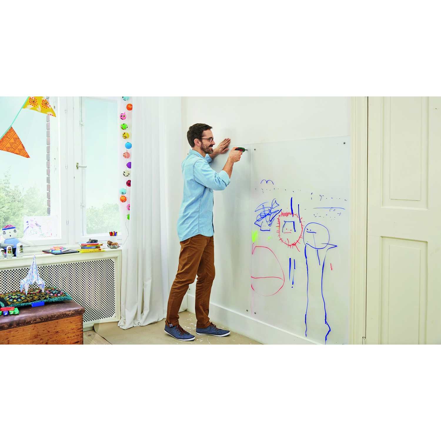 Uomo con camicia blu e pantaloni marroni attacca una lavagna a un muro con disegni colorati in una stanza.