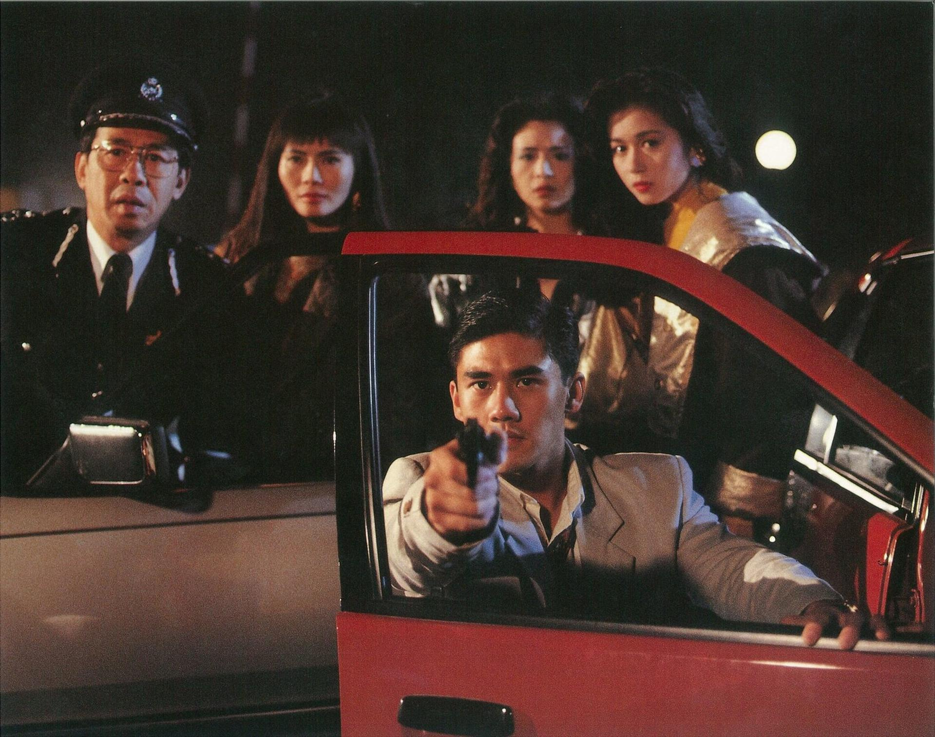 Ein Mann im Auto zielt mit einer Waffe. Andere, darunter ein Polizist, schauen zu. Rotes Auto, dunkler Hintergrund.