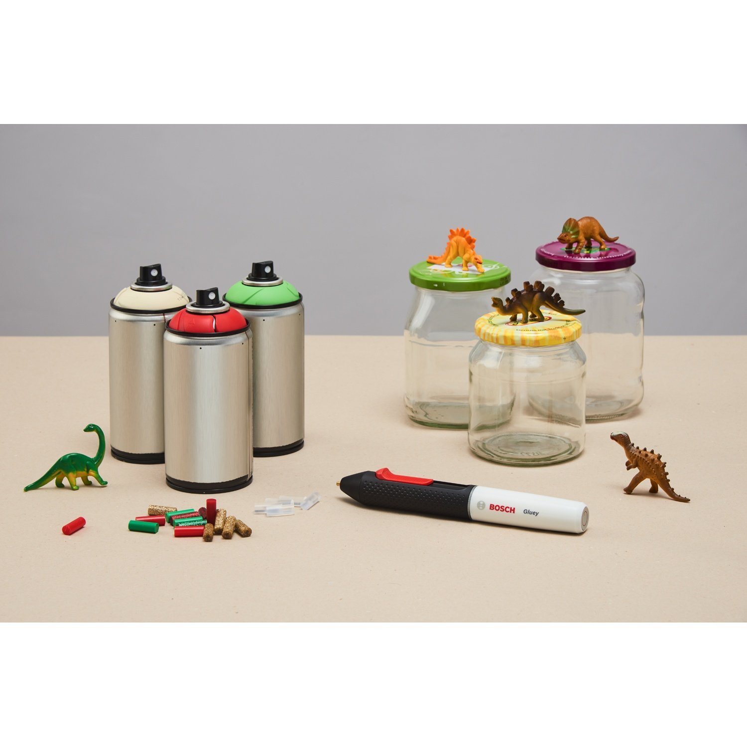 L'immagine mostra bombolette spray, barattoli con coperchi a dinosauro, dinosauri giocattolo, pistola per colla e stick di colla su una superficie marrone chiaro.