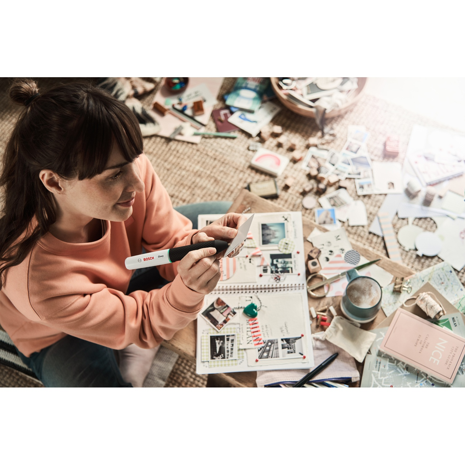 Una donna seduta su un tappeto tiene una penna sopra un album fotografico. Circondata da foto, mappe e una tazza di caffè.
