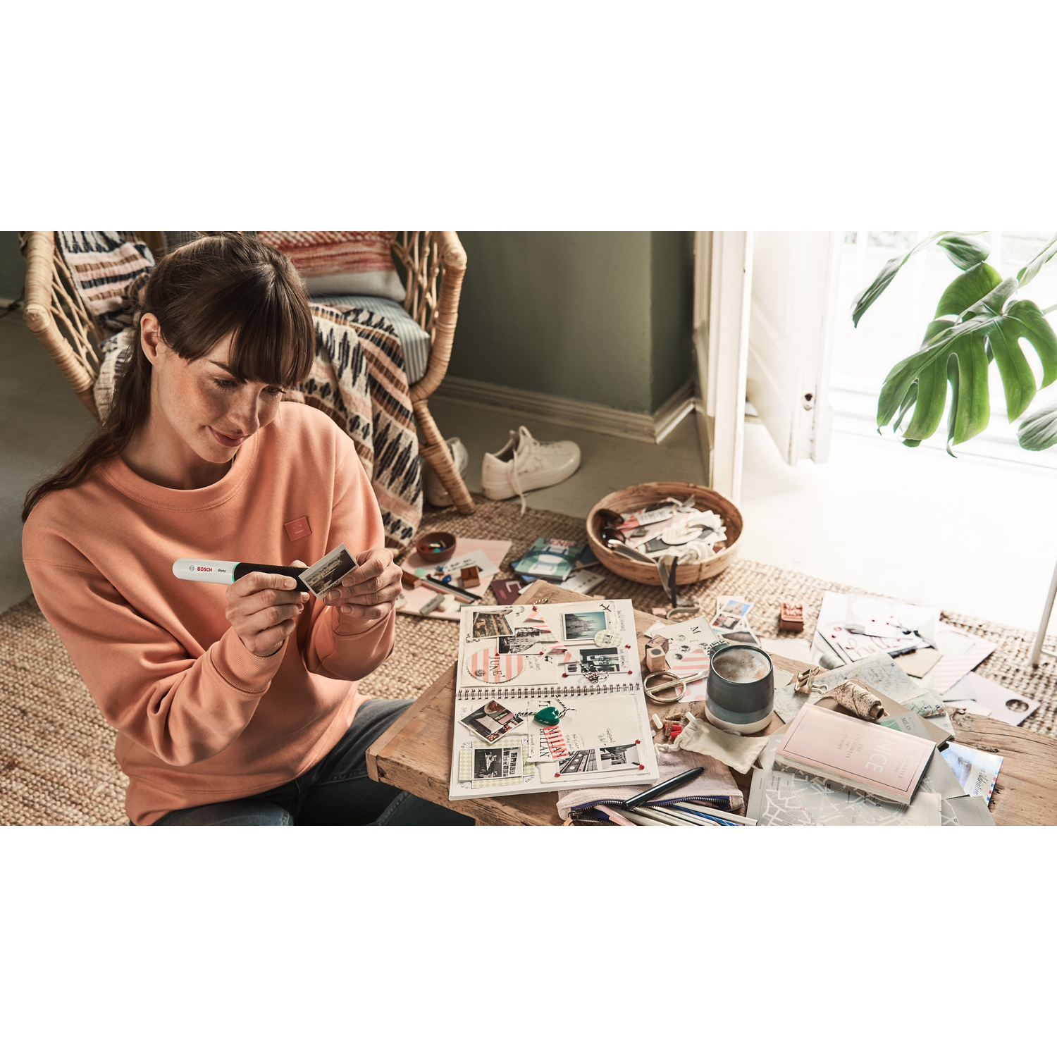 Una donna con un maglione color pesca è seduta a un tavolo di legno, facendo scrapbooking. Oggetti sparsi come foto, un diario e una pianta sono intorno.