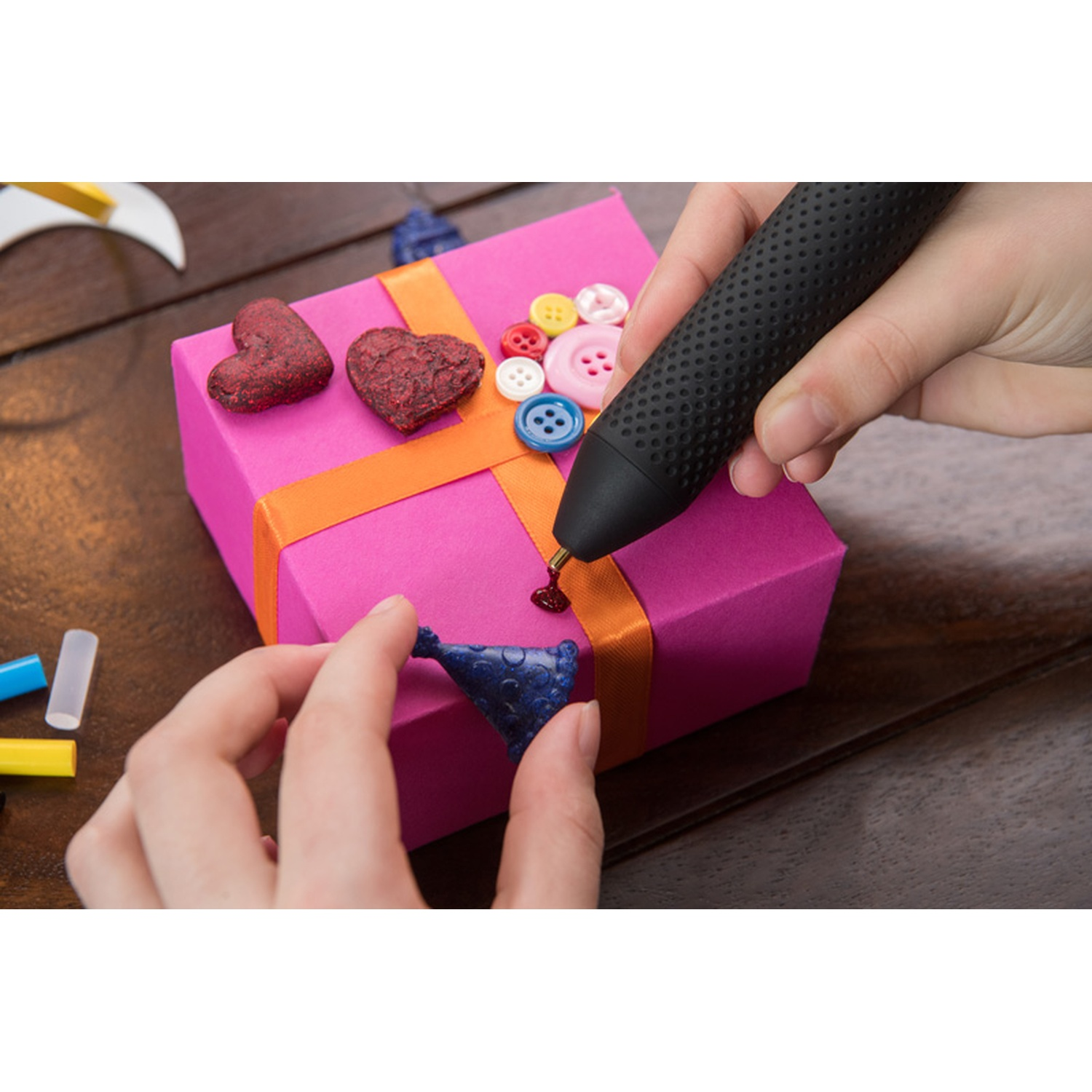 Una persona usa una penna nera per incollare una decorazione blu su una scatola regalo rosa con un nastro arancione. Ci sono bottoni e cuori.