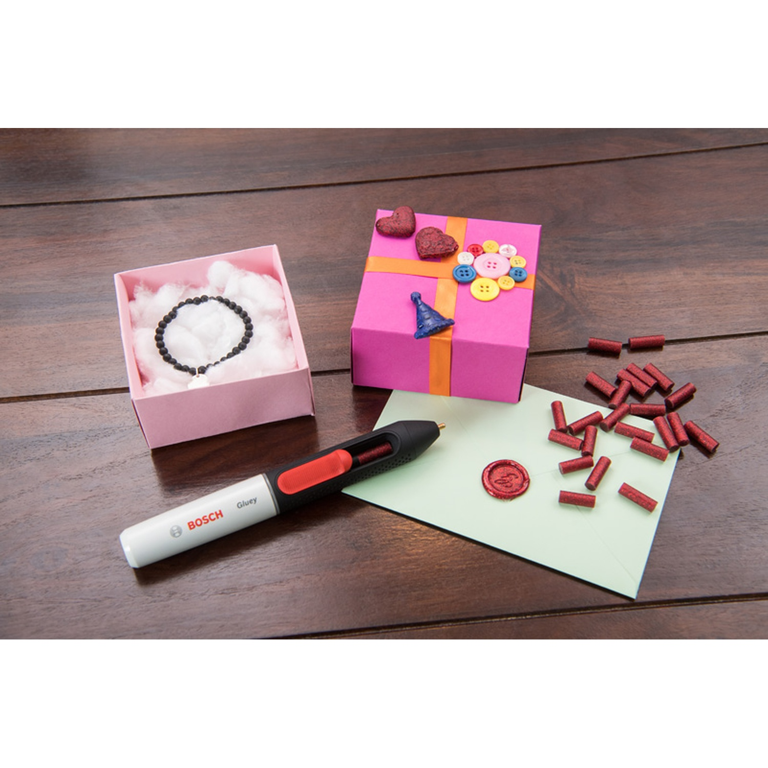 Scatola regalo rosa, braccialetto, pistola per colla, bastoncini di cera rossa, busta e scatola con braccialetto su superficie in legno.