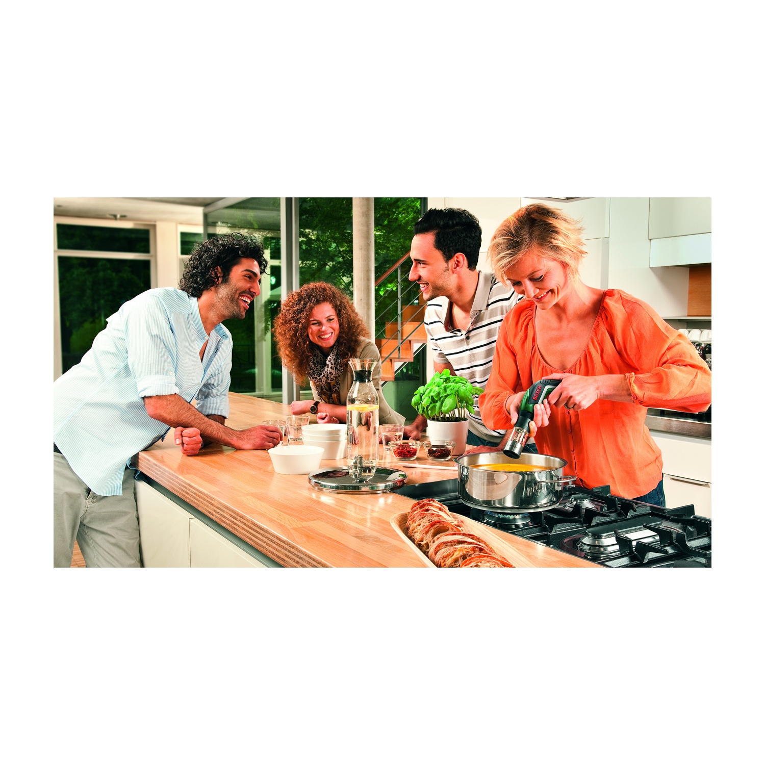 Quattro persone in cucina. Una donna con una camicia arancione cucina. Altri la guardano. C'è pane su un tagliere di legno.