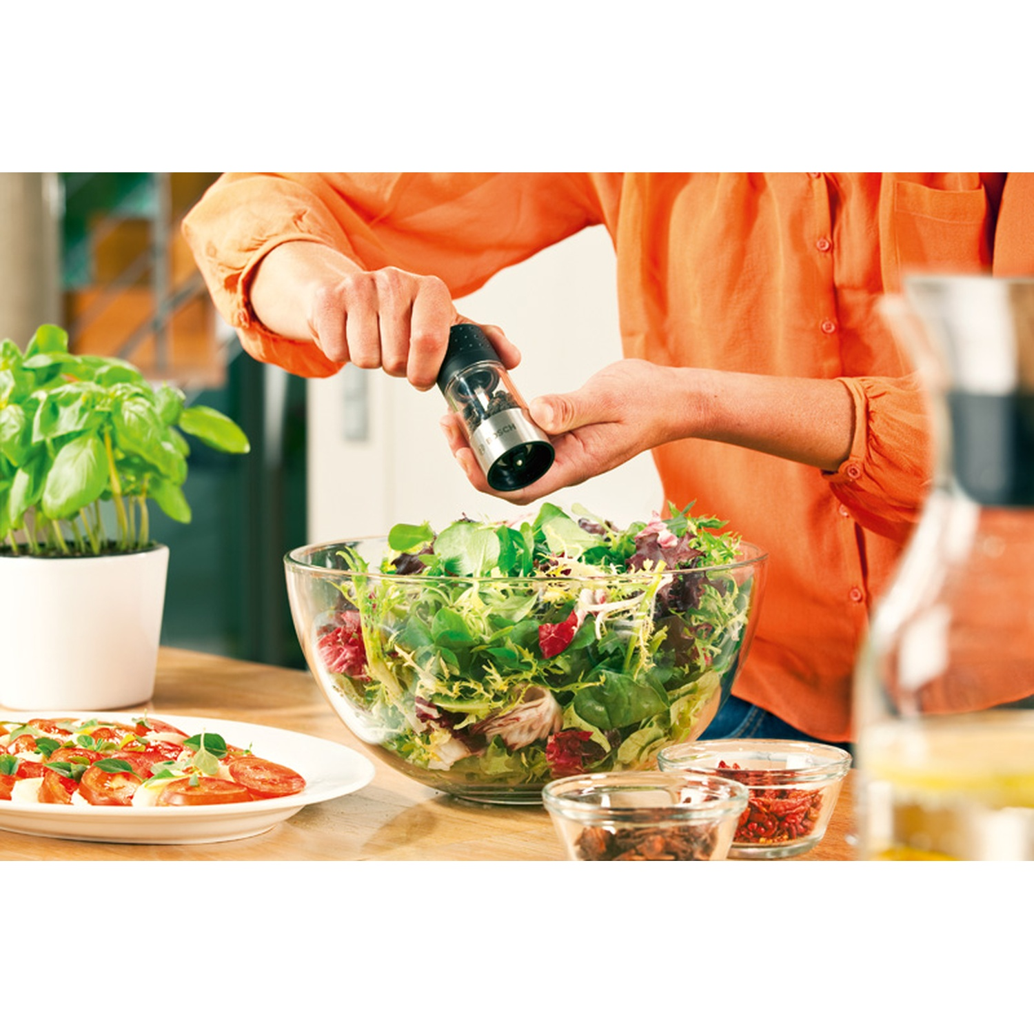 Una persona sta condendo un'insalata con un macinapepe. Una grande ciotola di vetro contiene l'insalata. Altri ingredienti e pianta di basilico sono presenti.