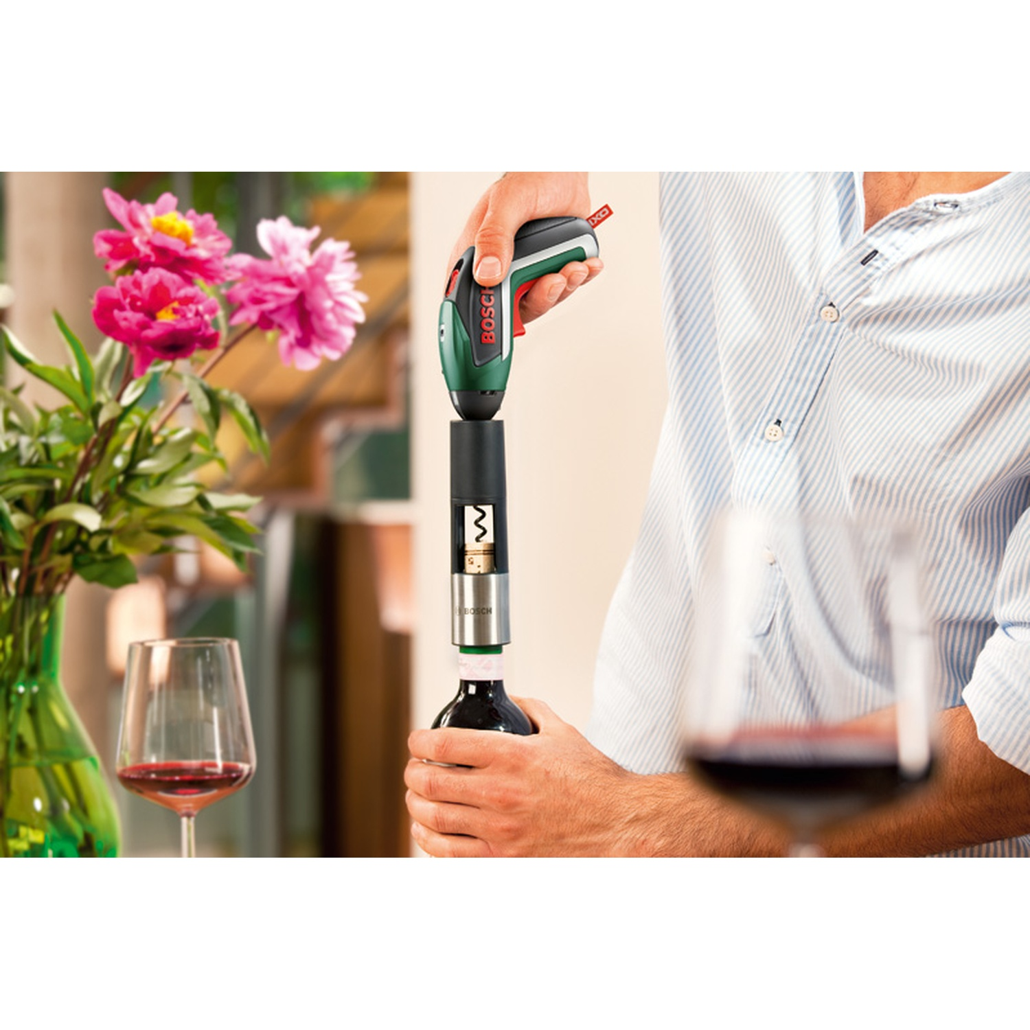 Un uomo usa un apribottiglie elettrico Bosch. Una bottiglia di vino viene aperta. Due bicchieri di vino e fiori sono sullo sfondo.