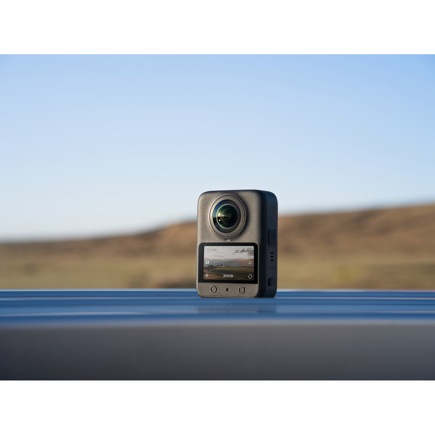 Een zwarte 360-graden camera staat op een blauw oppervlak met een scherm dat een landschap toont. Achtergrond: wazig veld en lucht.