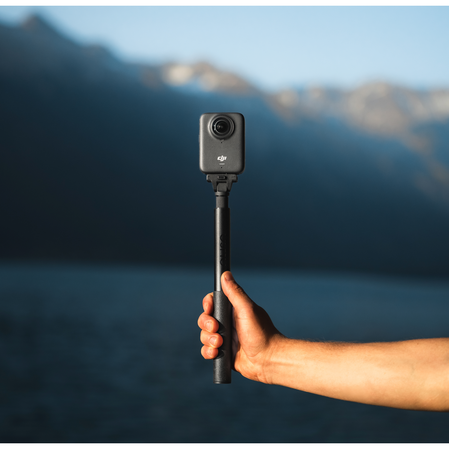 Une personne tient une caméra d'action noire sur une perche à selfie. Montagnes et eau en arrière-plan flou. Le ciel est bleu.