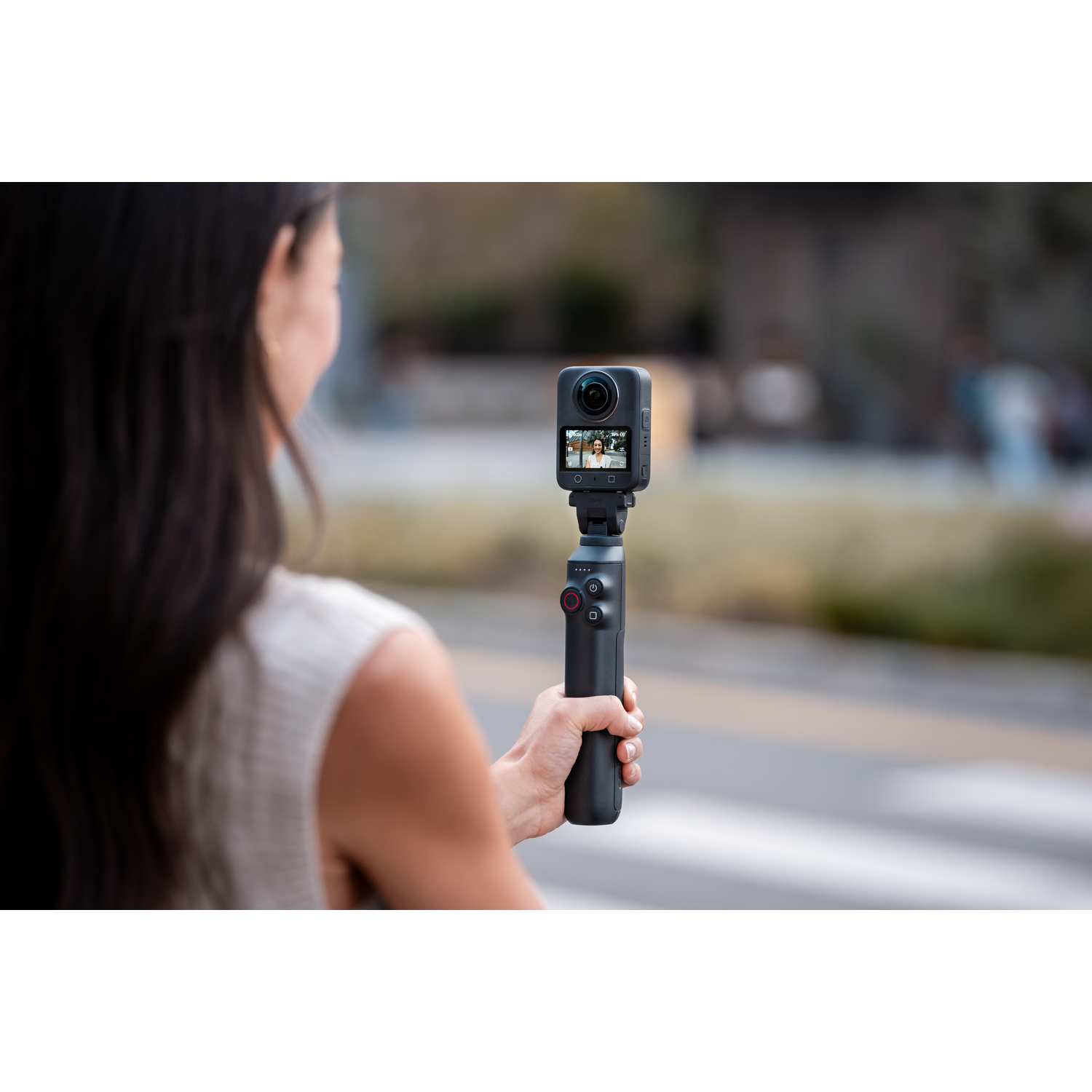 Woman holding a black camera on a stick, recording video outdoors. Background includes a blurred street and greenery.