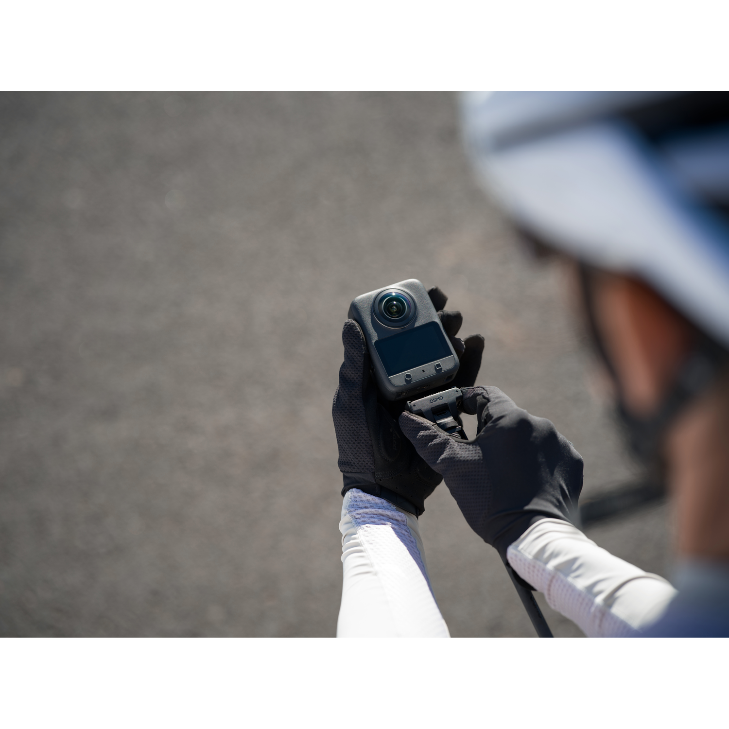 A person in cycling gear holds a small camera. Black gloves and a white sleeve are visible. A gray road fills the background.
