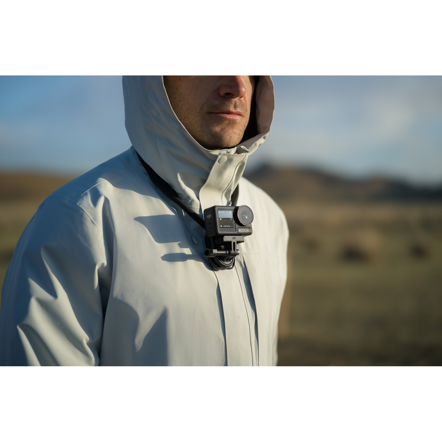 Man wearing a light gray hooded jacket, with a camera hanging around the neck. Background has a blurry landscape.