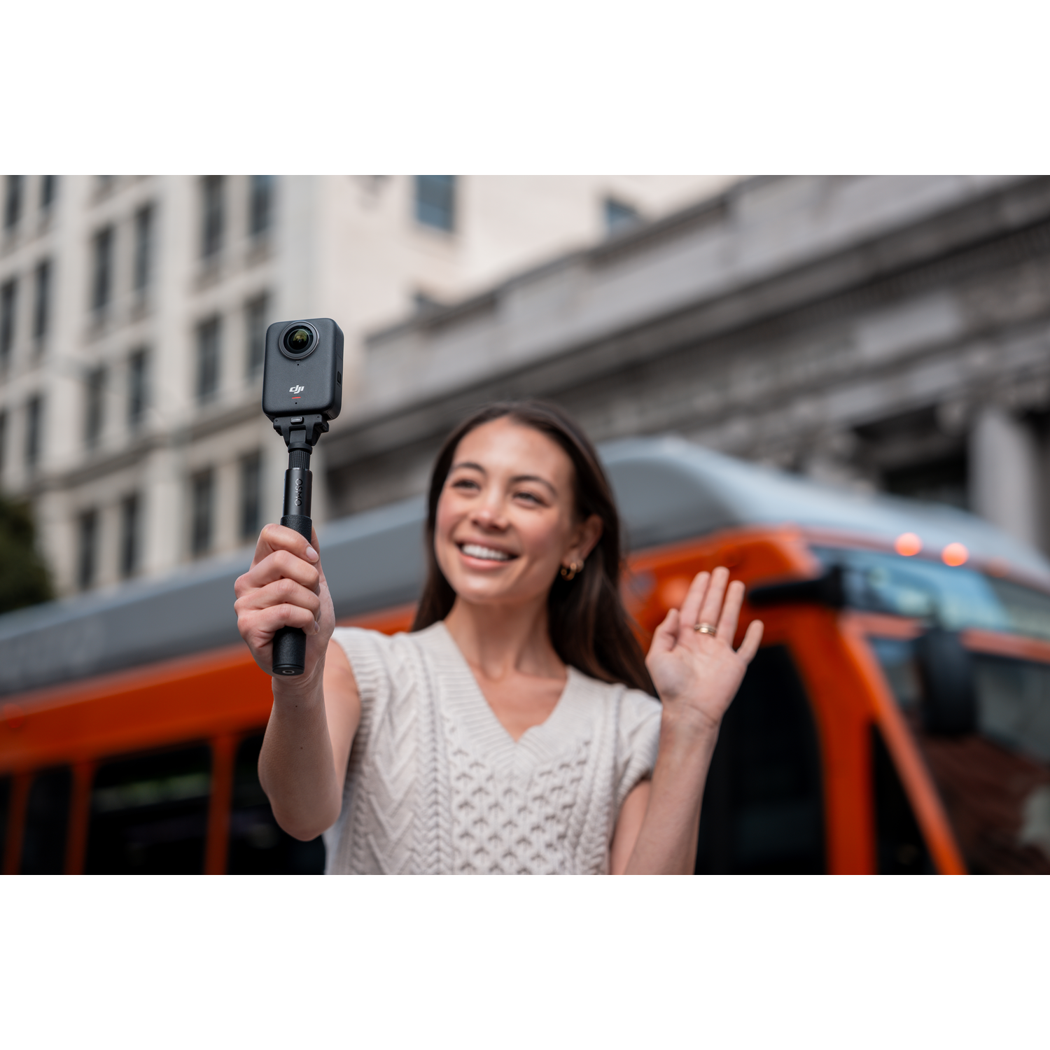 Woman holding camera, waving in front of an orange bus and buildings. She is smiling, wearing a white sweater. The camera is black.