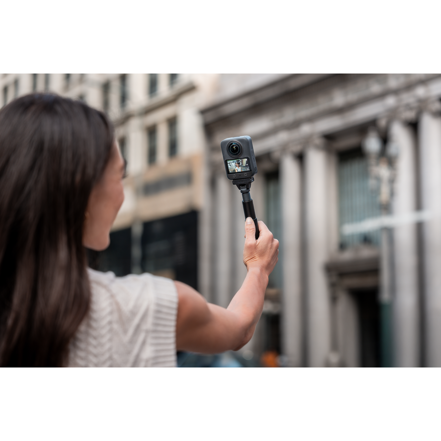 A woman holds a 360 camera, taking a selfie in front of a building. The camera displays a live view.