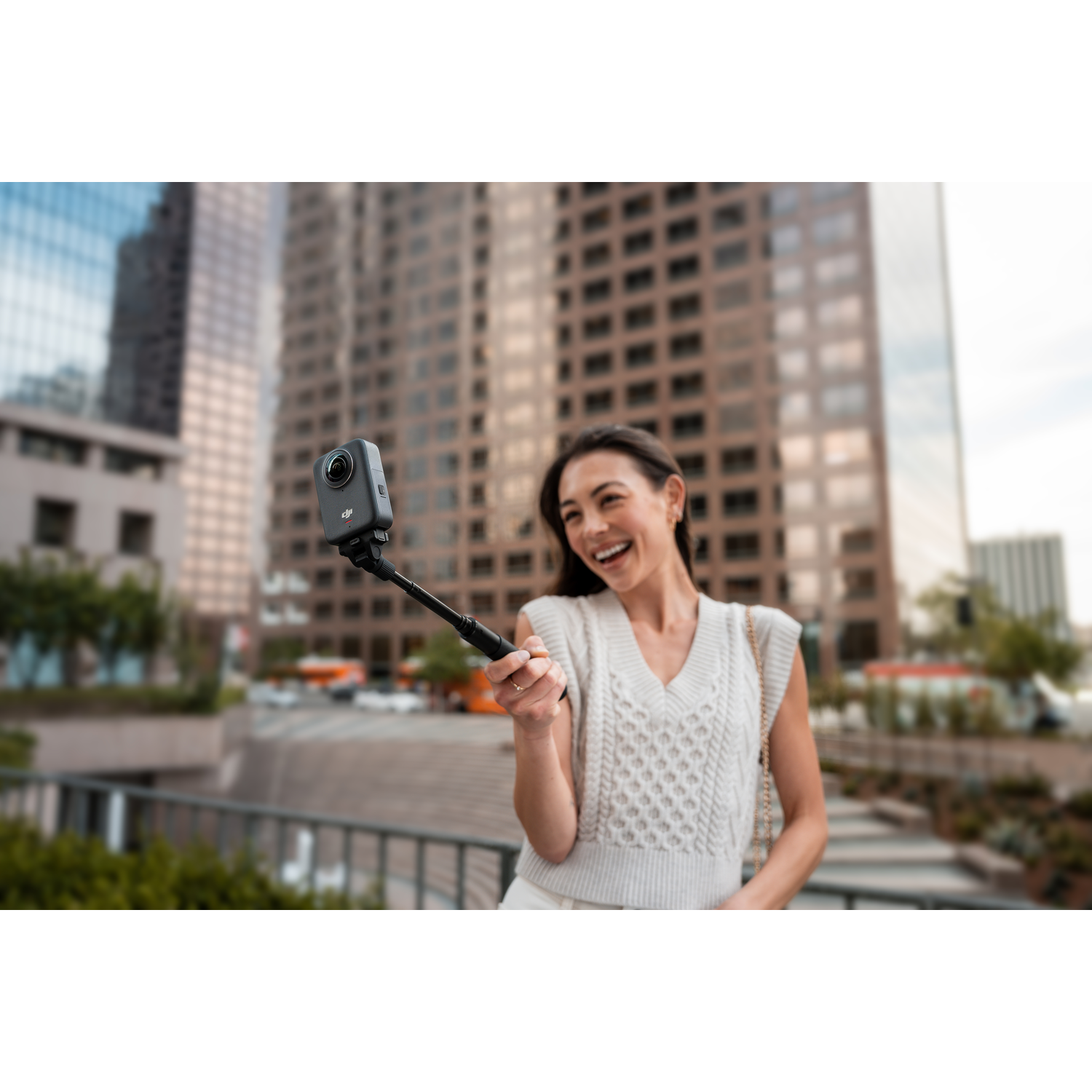 Woman holding a black camera on a selfie stick, smiling in front of city buildings.