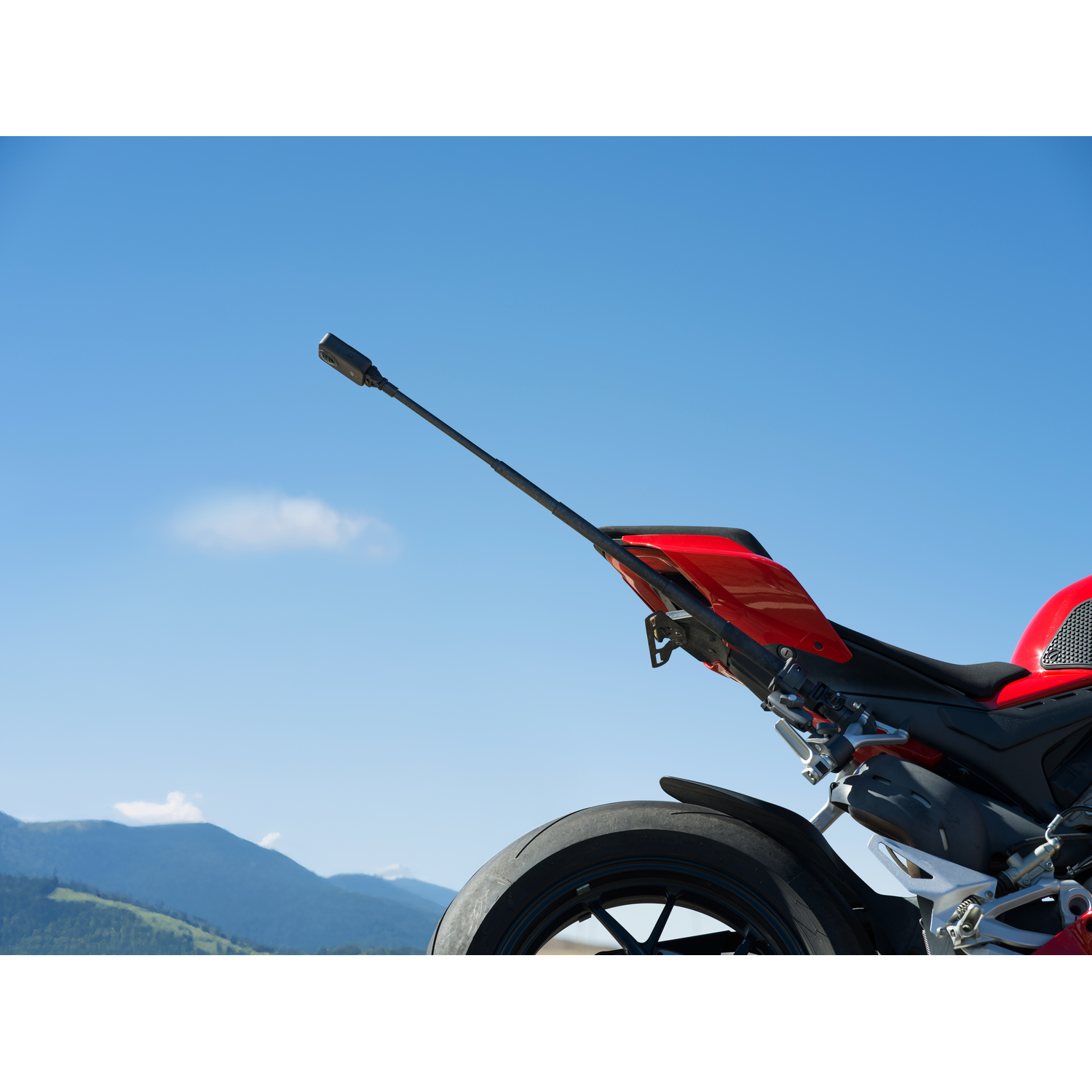 Moto rouge avec pneu noir et caméra sur un poteau contre ciel bleu et montagnes en arrière-plan.