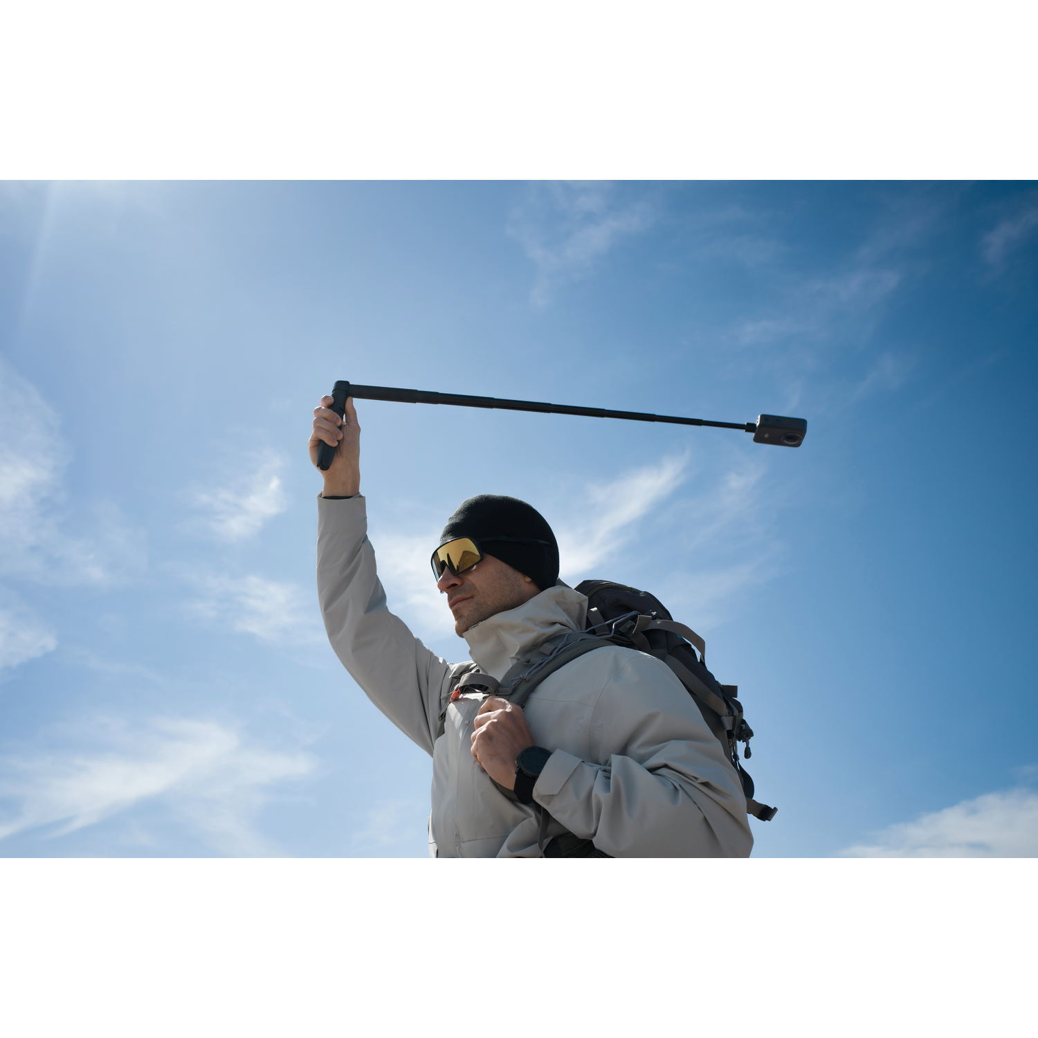 Man in sunglasses and hat holds a camera on a stick against a blue sky with white clouds. He wears a backpack.