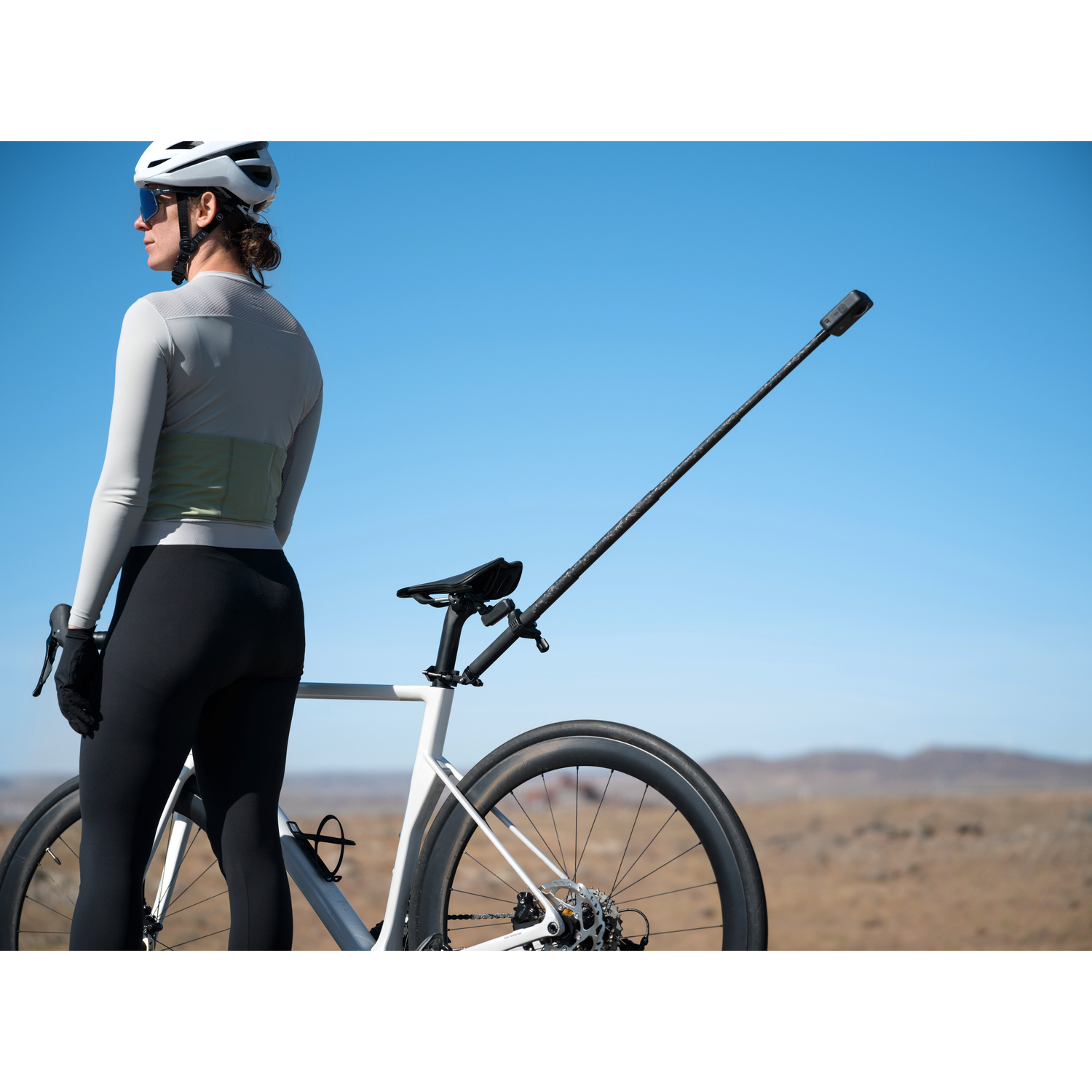 Cyclist with bike, helmet, and sunglasses stands against a blue sky. The bike has a camera attached. Background is a desert.