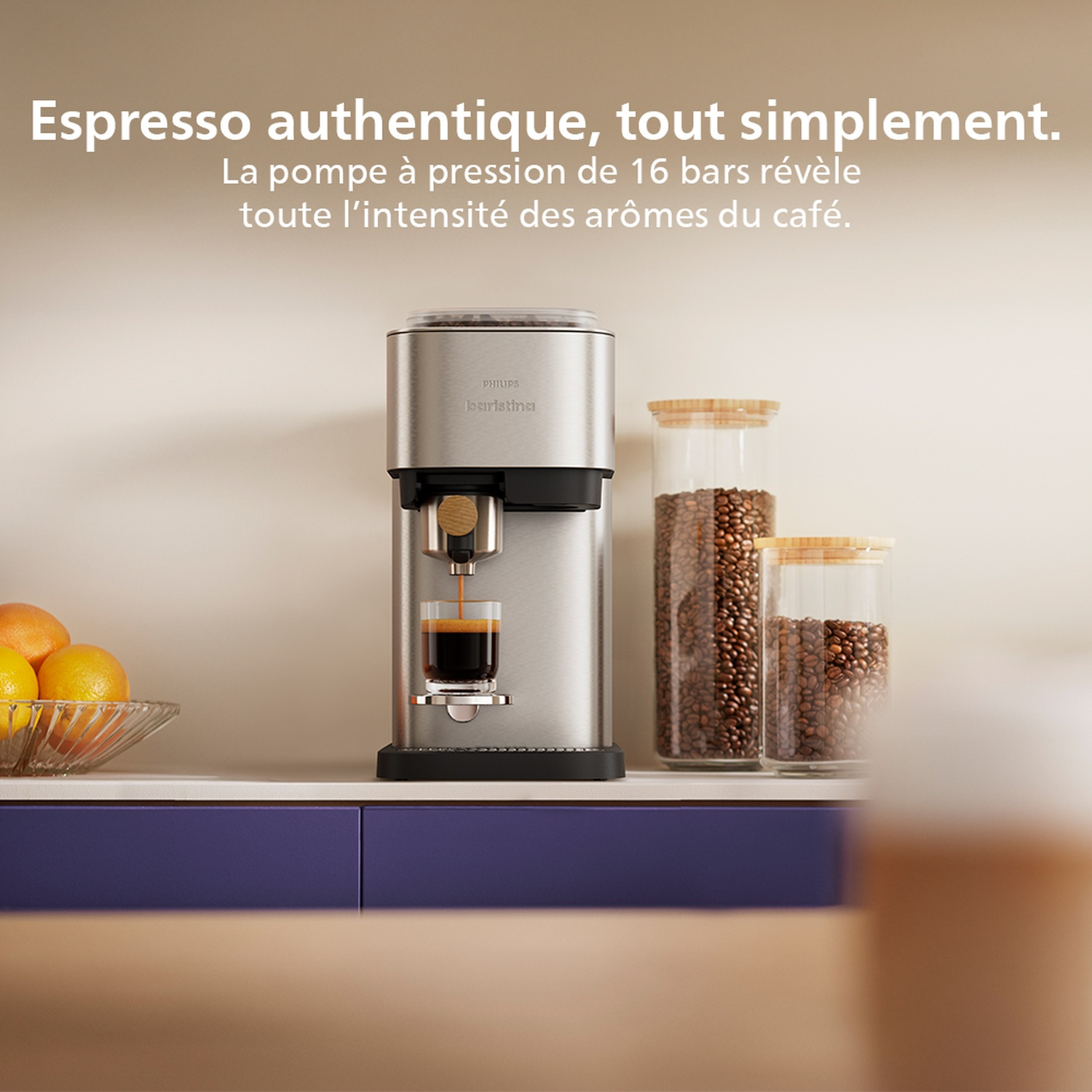 Machine à expresso argentée versant du café dans un verre. Grains de café dans des bocaux, oranges dans un bol, sur un comptoir blanc avec des placards bleus.