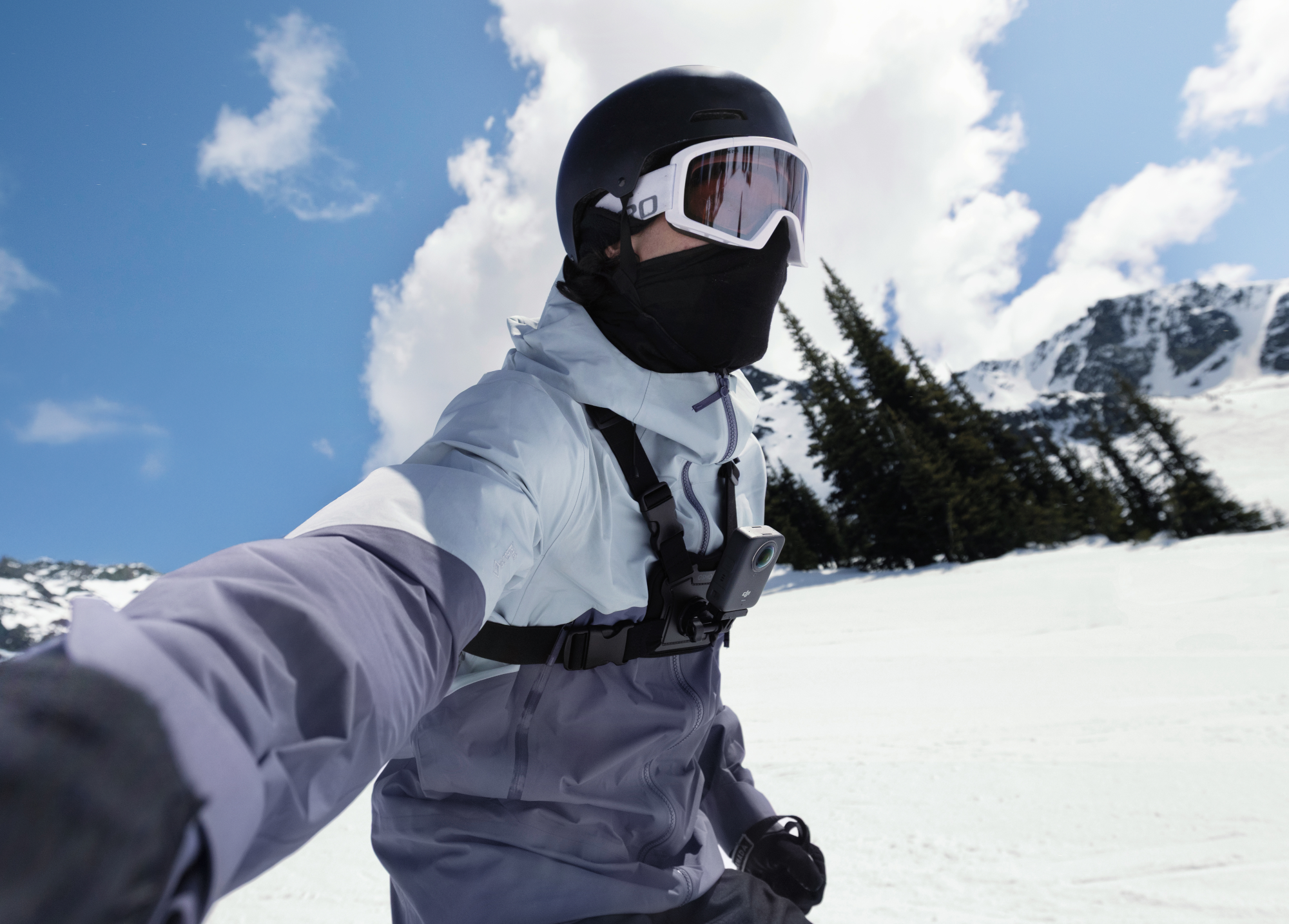 Skifahrer in schwarzem Helm, Brille und grauer Jacke auf schneebedeckter Piste. Blauer Himmel und Berg mit Bäumen im Hintergrund.