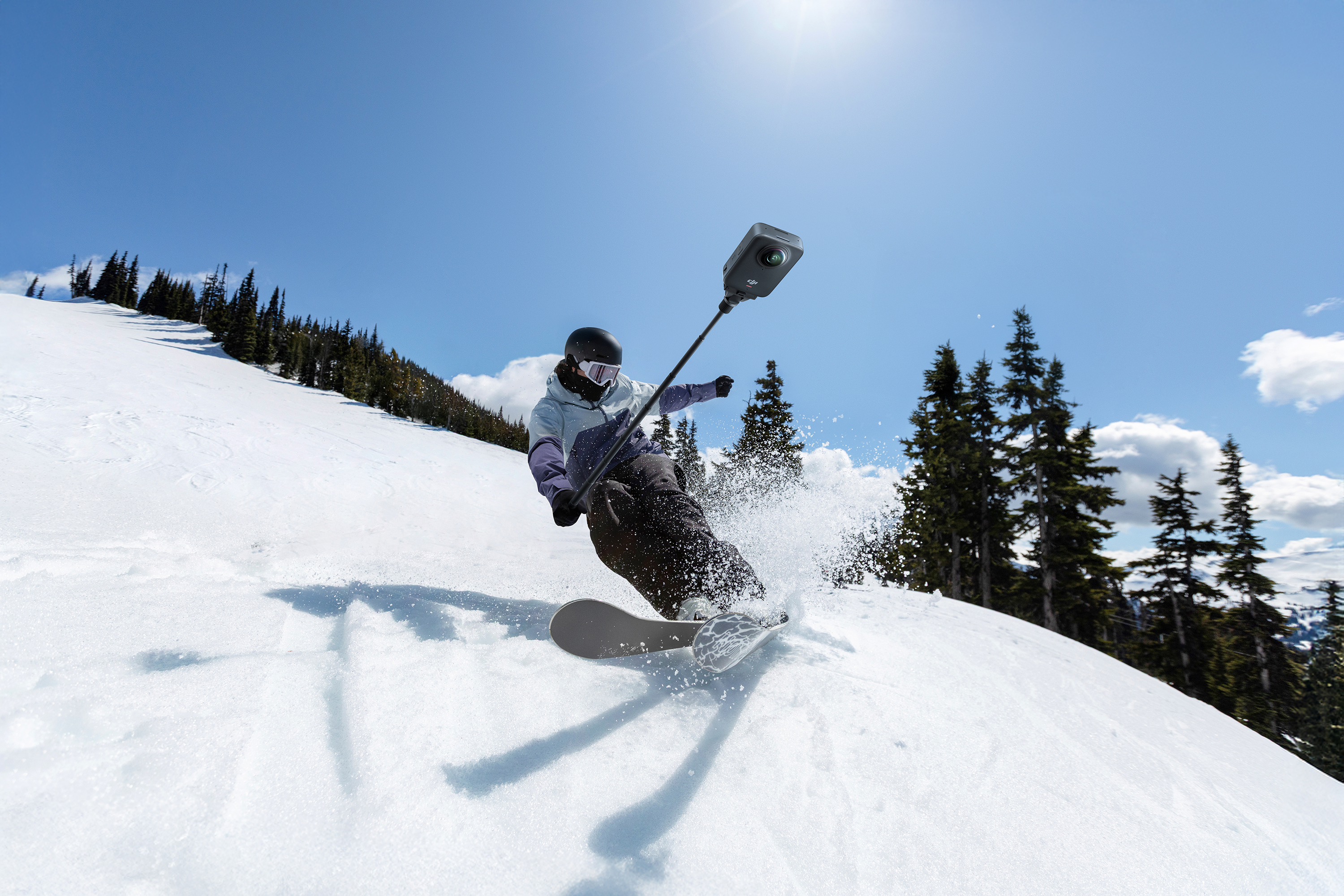 Snowboardos a havas lejtőn kamerával, kék égbolttal és fákkal. A nap süt. Hópermet a deszkáról.
