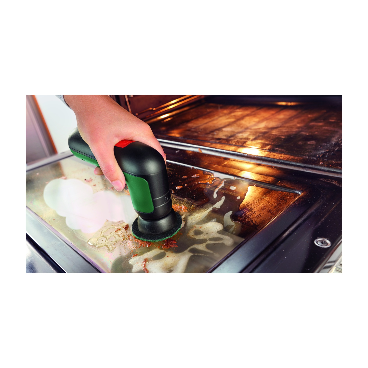 Una mano usa uno strumento di pulizia verde e nero per strofinare una porta del forno sporca. Schiuma saponosa e residui sono visibili.