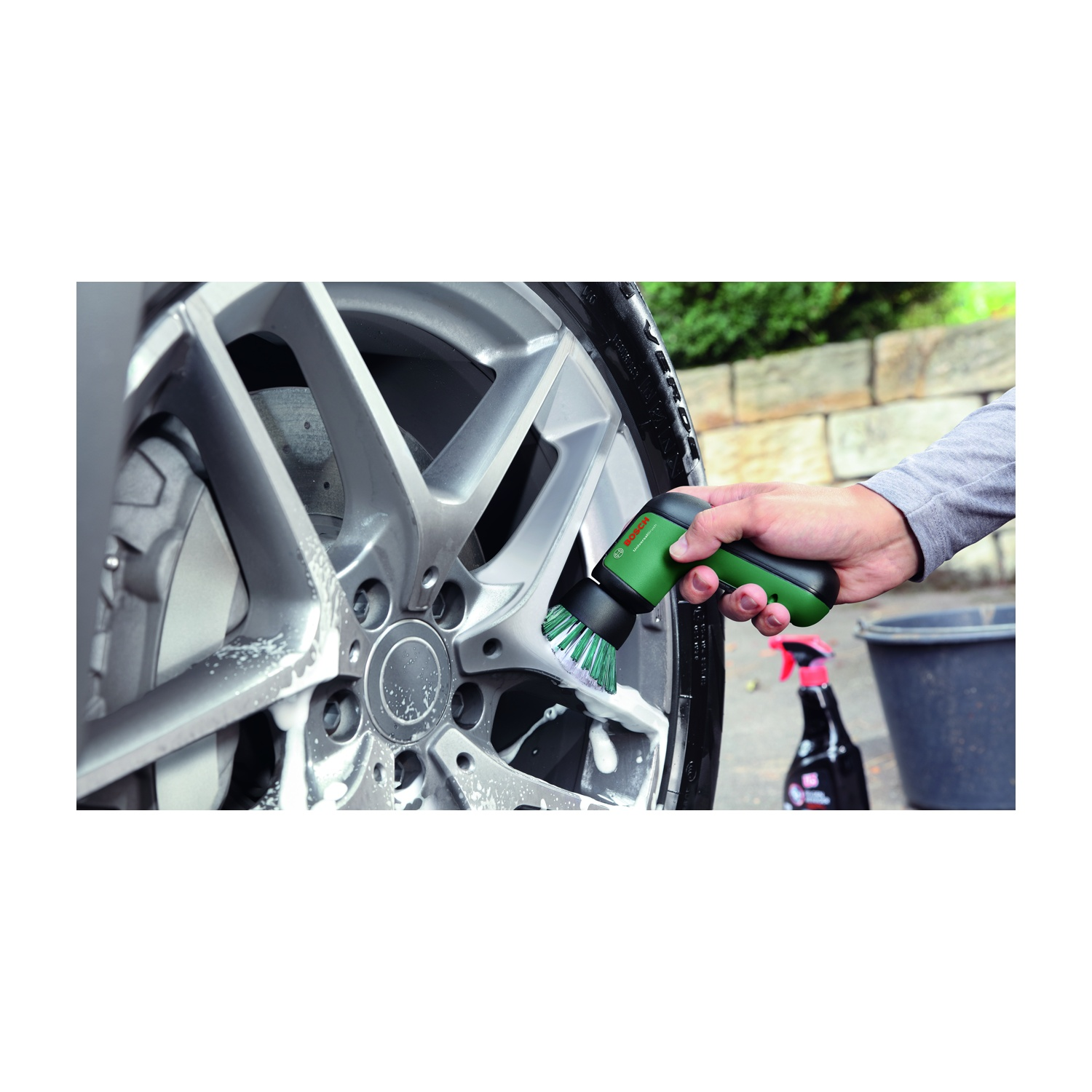Una persona pulisce una ruota di un'auto con una spazzola verde e nera. Schiuma di sapone copre la ruota. Uno spruzzatore e un secchio sono dietro.