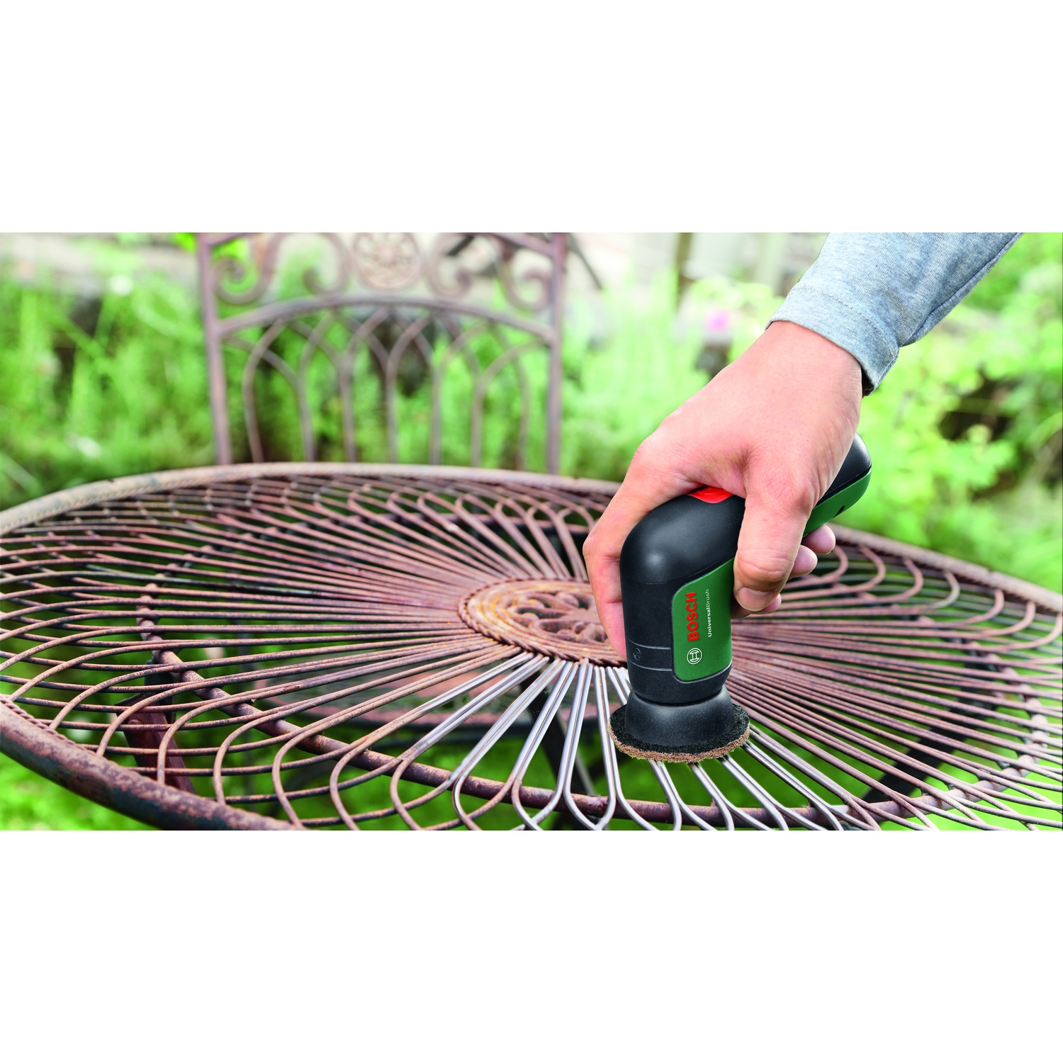 Una mano usa uno strumento Bosch per pulire un tavolo di metallo arrugginito all'aperto. Una sedia in ferro battuto e fogliame verde sullo sfondo.