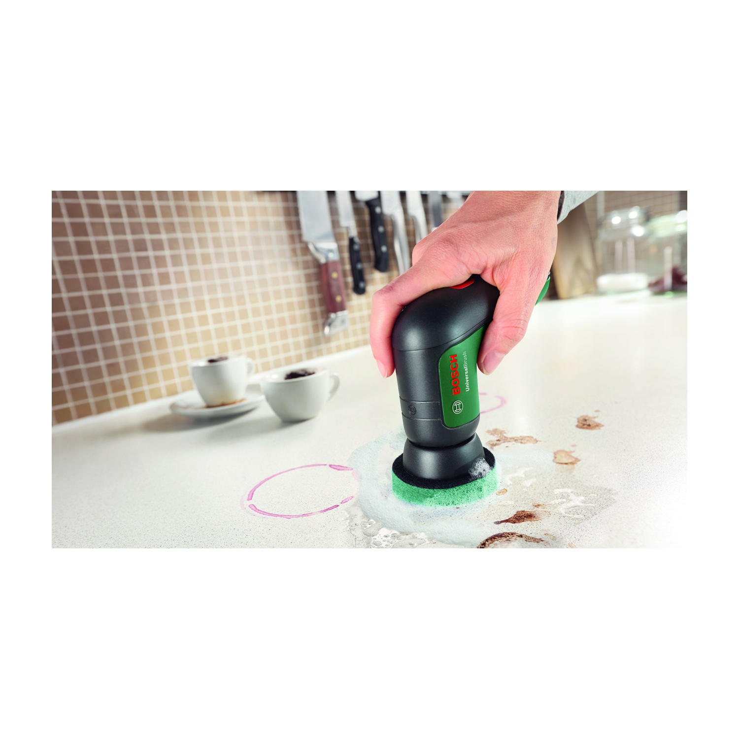 Una mano tiene una spazzola per la pulizia Bosch, strofinando un piano di lavoro bianco. Macchie di caffè e un anello rosa segnano la superficie. Tazze e coltelli sullo sfondo.