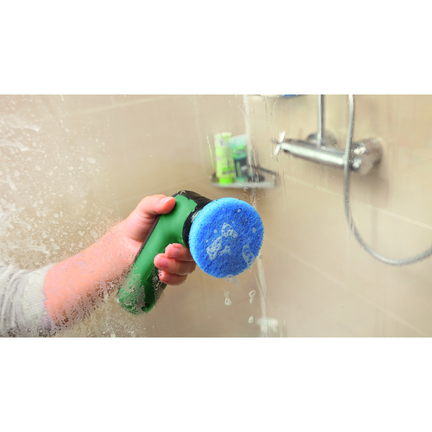 Una mano che tiene uno strumento per la pulizia verde con una spugna blu che pulisce un vetro della doccia. Sapone e soffione sono visibili.