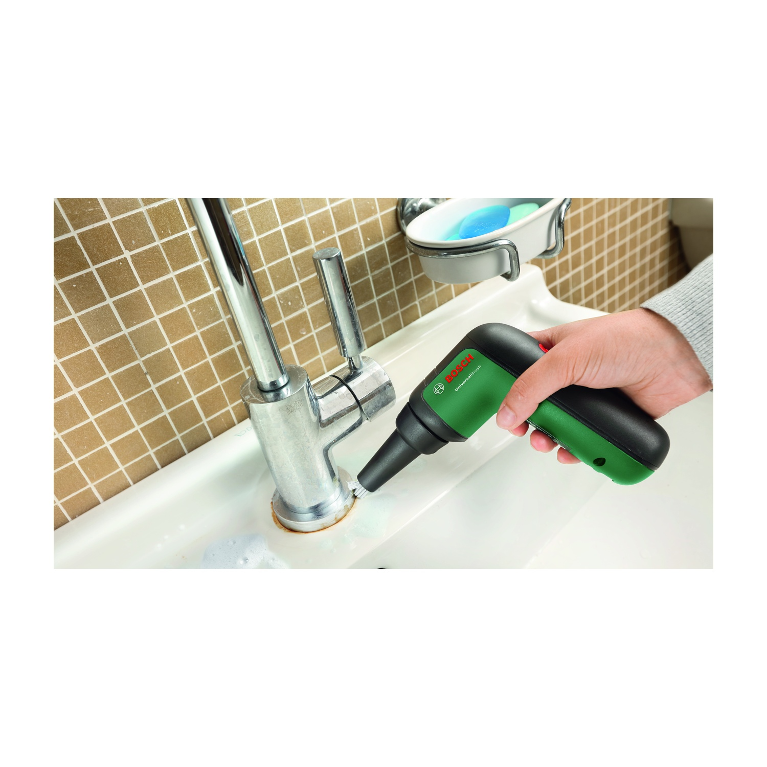 Una mano che tiene un elettroutensile Bosch verde e nero che pulisce un rubinetto. C'è un lavandino bianco, un portasapone e un muro piastrellato.