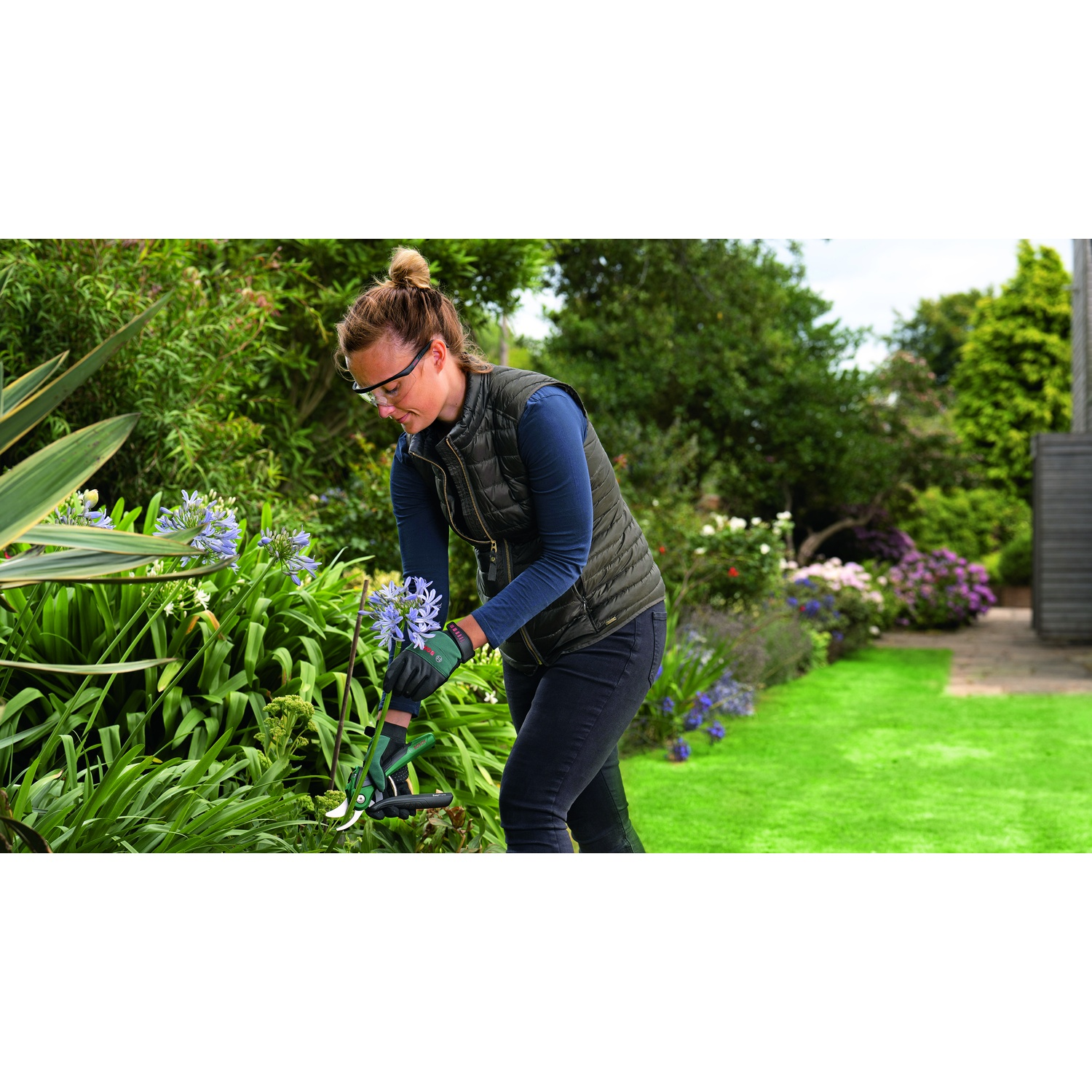 Donna in giardino pota fiori blu con cesoie verdi. Indossa occhiali, gilet e guanti. Erba verde e cespugli sullo sfondo.