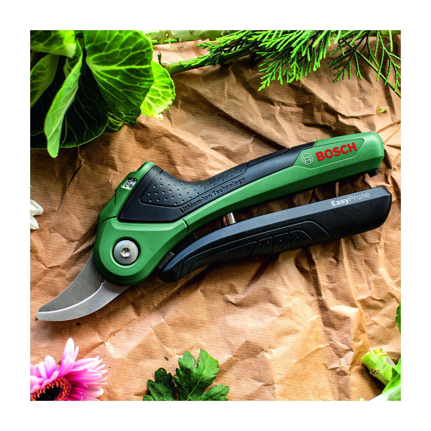Utensile verde Bosch EasyPrune su carta marrone con foglie e fiori. L'utensile è parzialmente aperto. Cavolo e parti verdi di piante lo circondano.