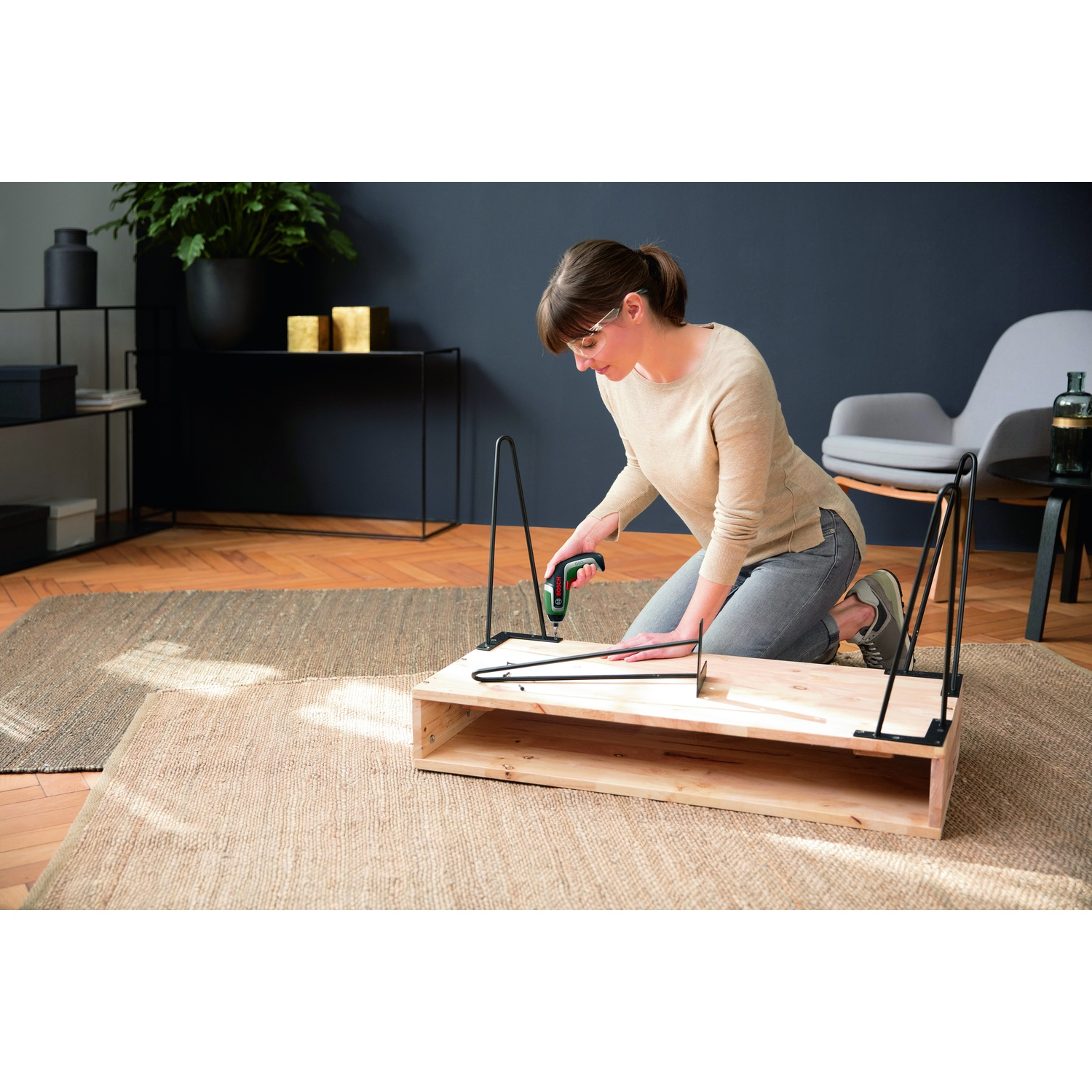 Una donna assembla un tavolo di legno, usando un trapano elettrico. È in ginocchio su un tappeto. Sono attaccate gambe nere.