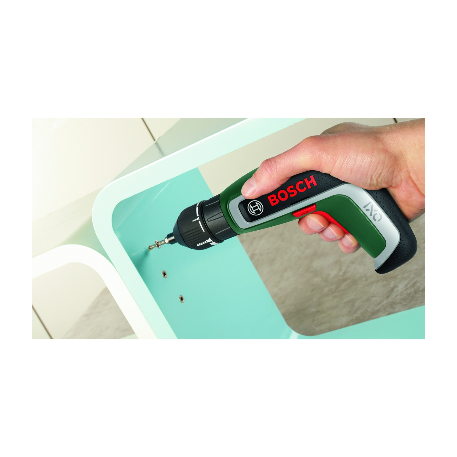 Una mano tiene un cacciavite a batteria Bosch IXO verde e nero. Sta avvitando in una superficie curva blu chiaro, con fori per viti visibili.