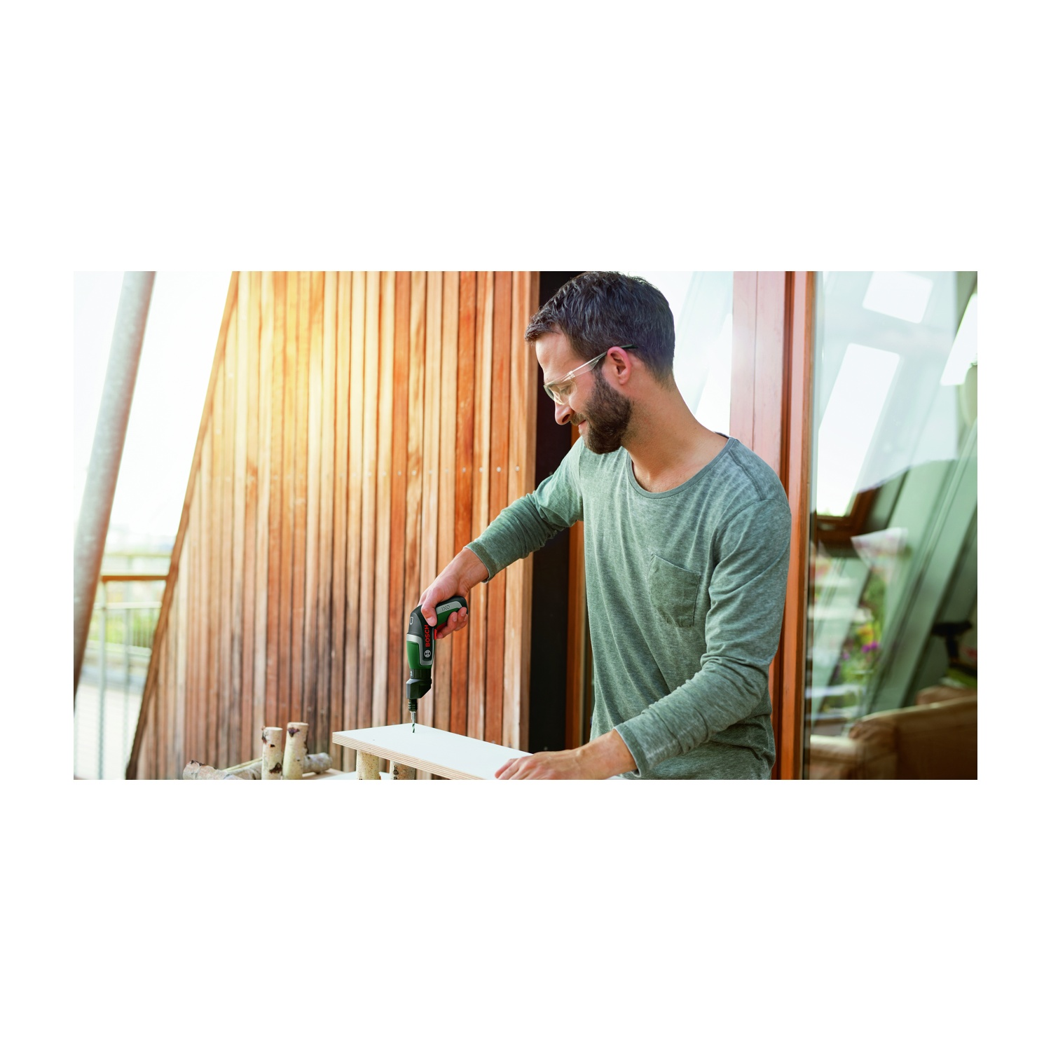 Uomo usa un trapano su una tavola di legno. Indossa occhiali e camicia verde. Sfondo con parete in legno e vetro.