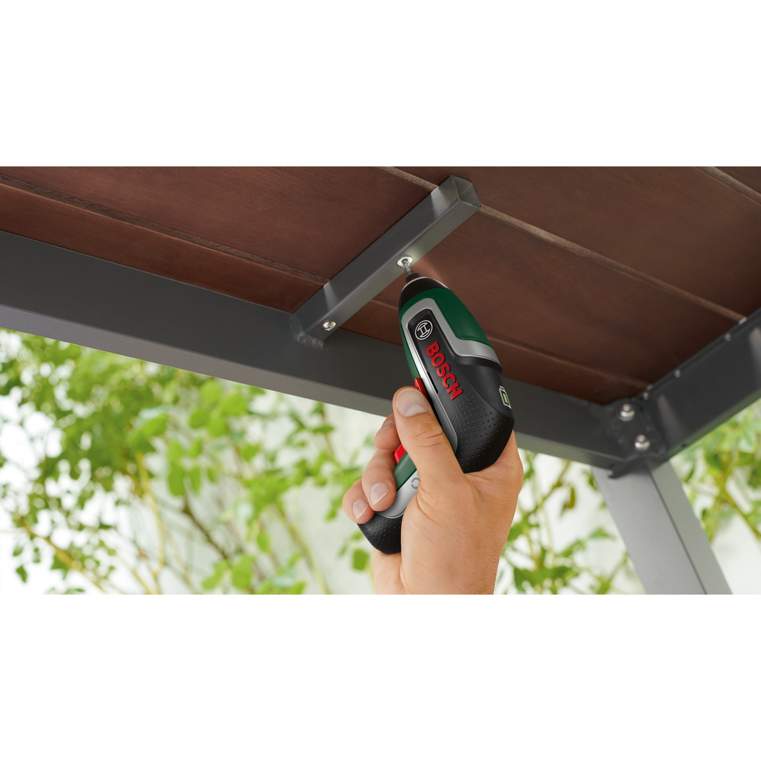 Mano che usa un avvitatore elettrico Bosch per stringere una vite in una struttura di legno. Fogliame verde sullo sfondo.