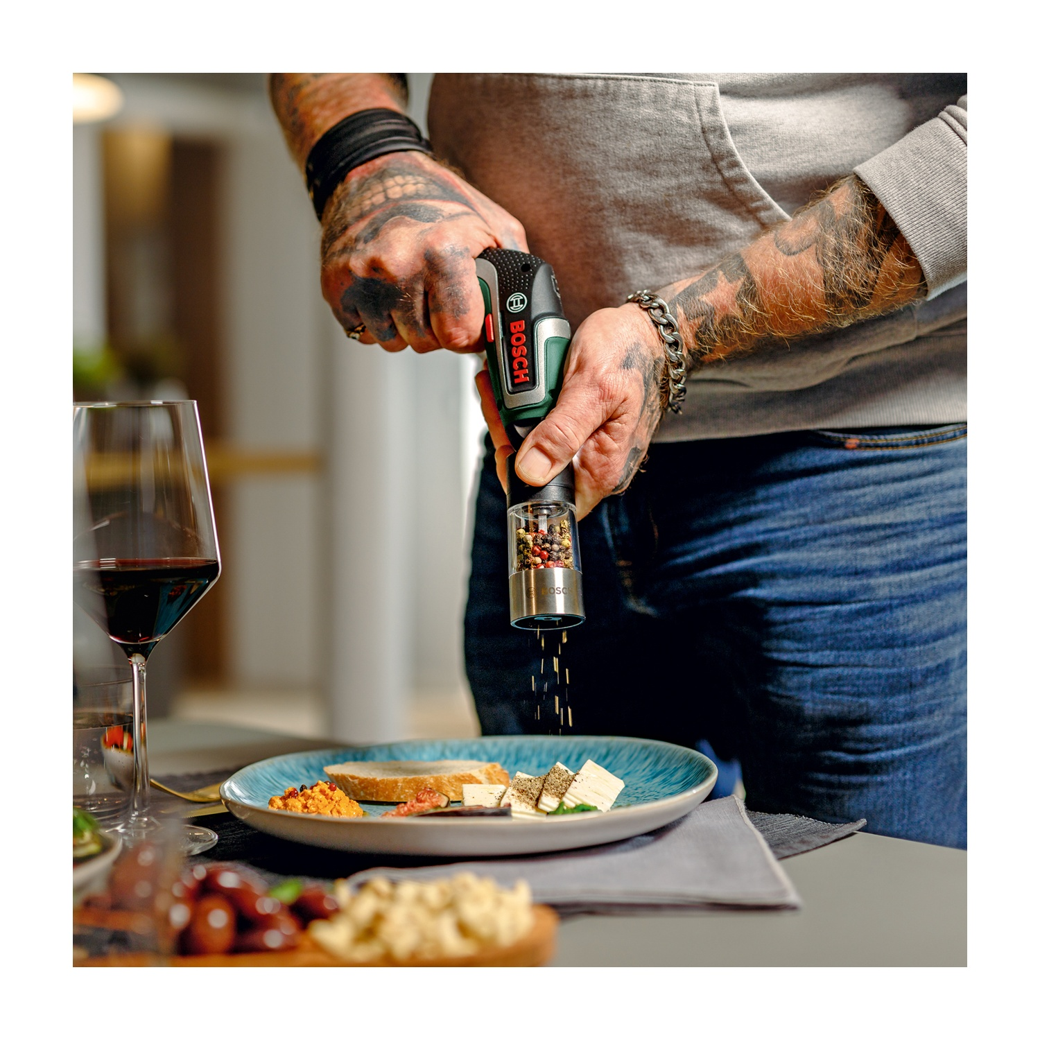 Una persona tatuata macina pepe sul cibo. Un piatto con formaggio e pane è su un tavolo con vino.